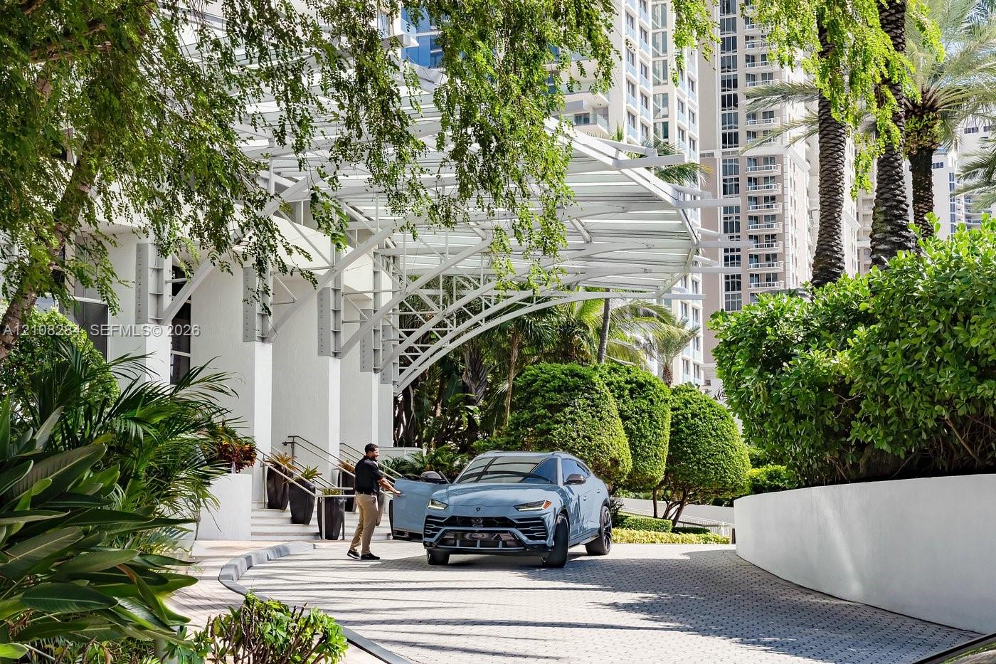 Porte-cochere entrance featuring white structural steel framing, paved circular driveway, and mature perimeter landscaping