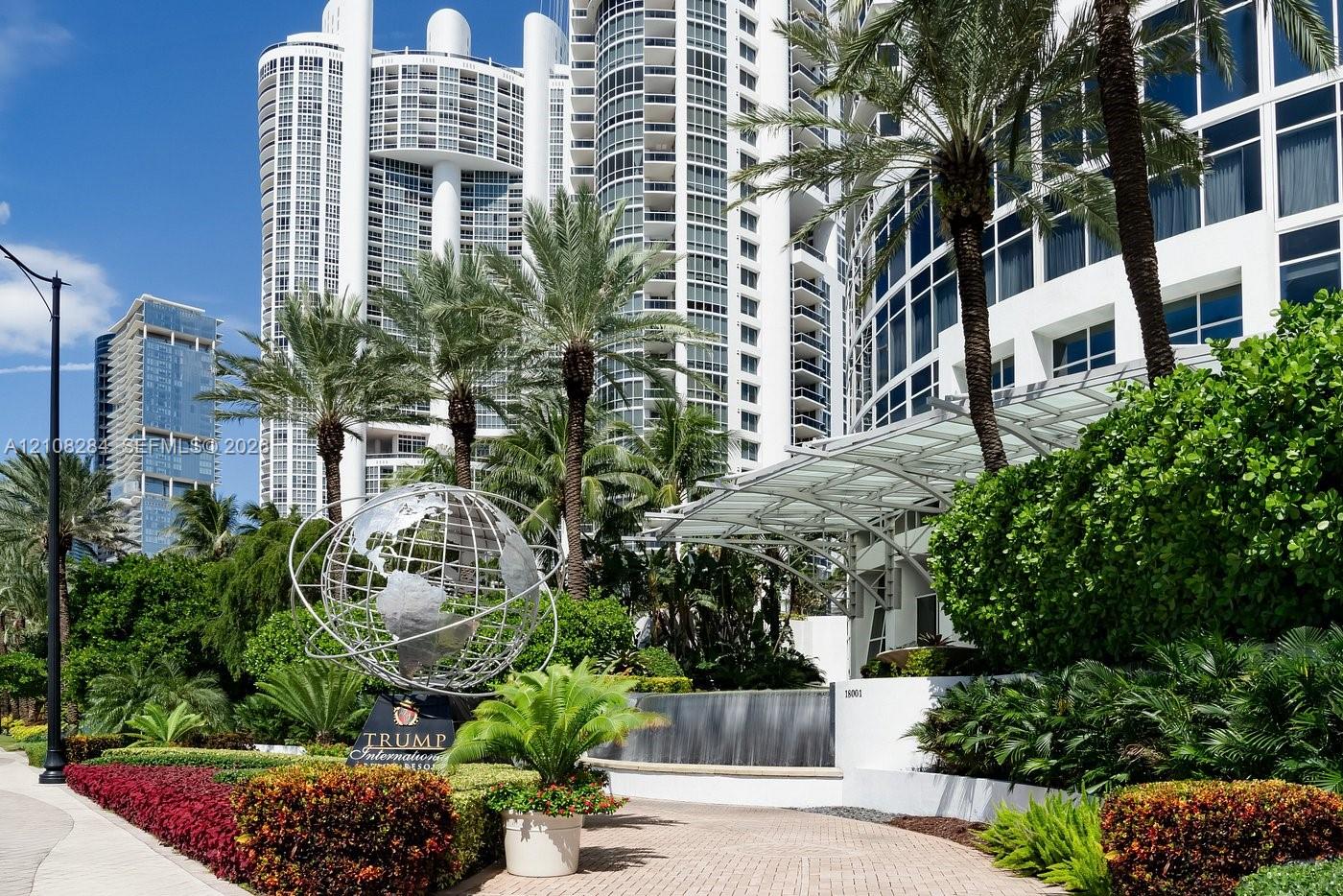 High-rise building exterior featuring a white curved facade, metal-frame porte-cochre, decorative metal globe sculpture, tiered water feature, and mature tropical landscaping