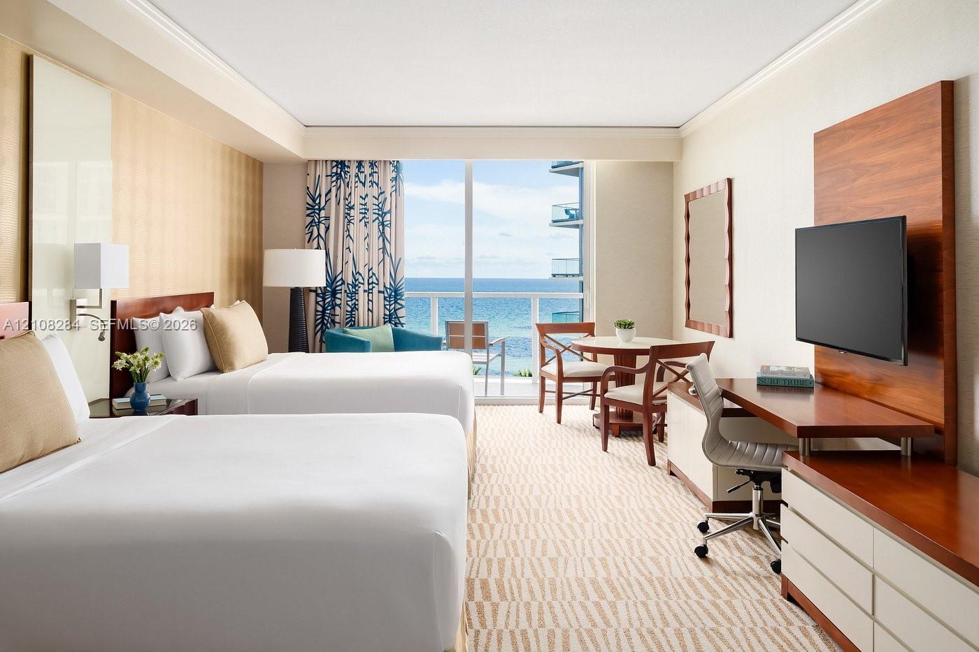 Floor-to-ceiling glass sliding doors opening to an exterior balcony, ocean backdrop, built-in wood-finish media console, integrated desk workspace, and recessed tray ceiling