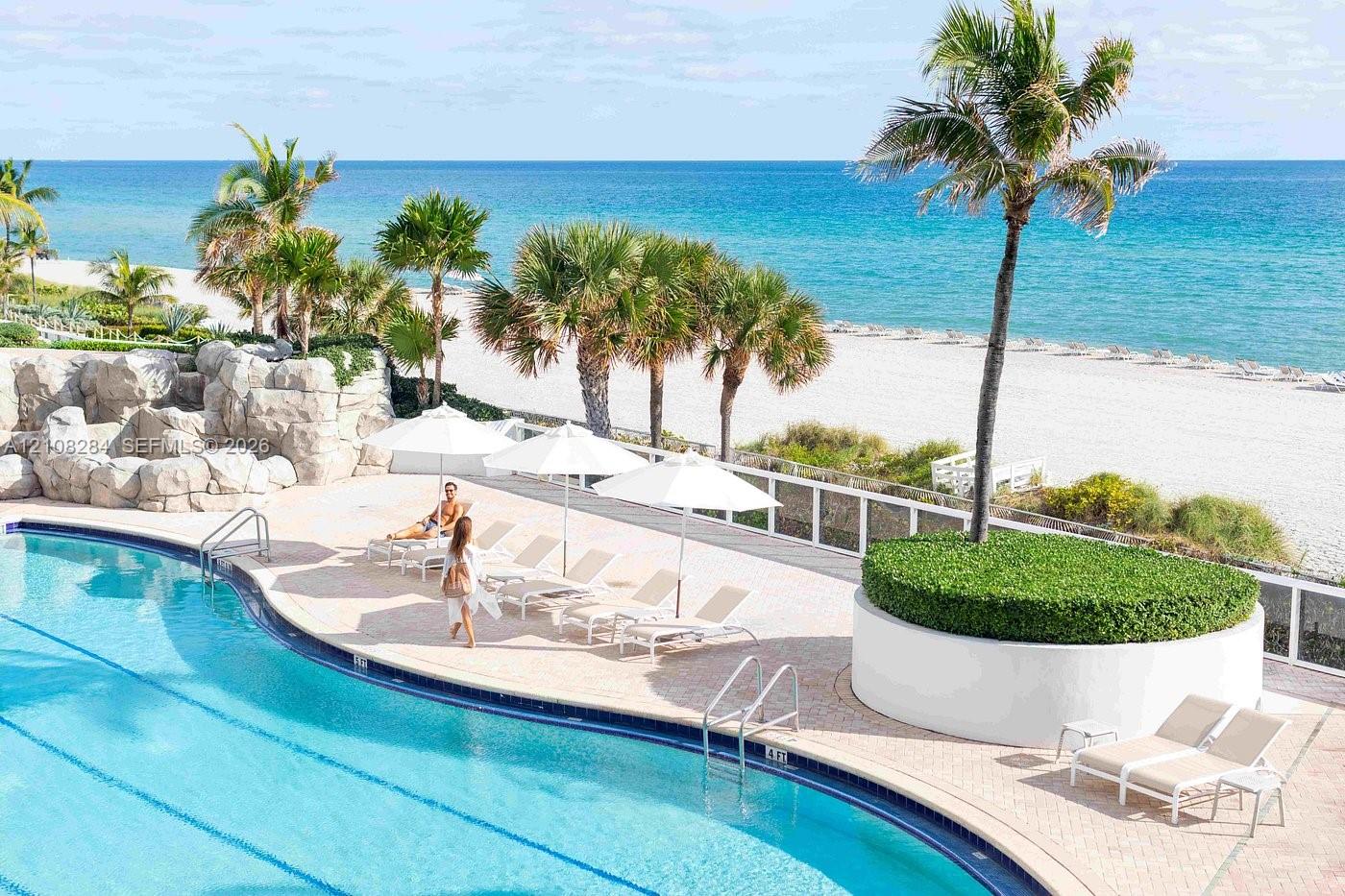 Curved swimming pool featuring a stone-faced grotto, expansive paver pool deck, elevated circular planter with mature palm tree, and white perimeter fencing