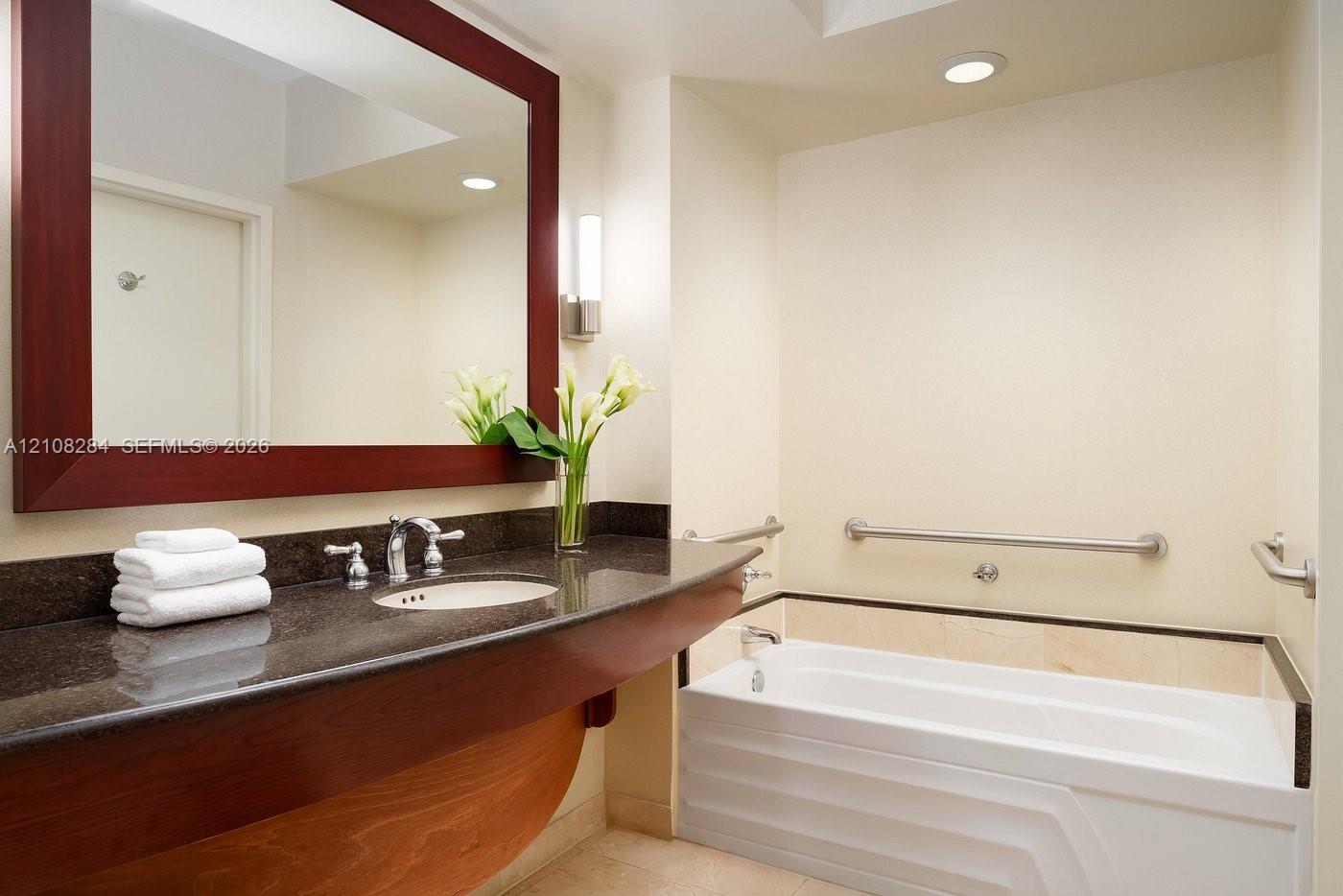 Floating wood vanity with dark stone countertop, integrated sink, large framed mirror, and bathtub with wall-mounted grab bars