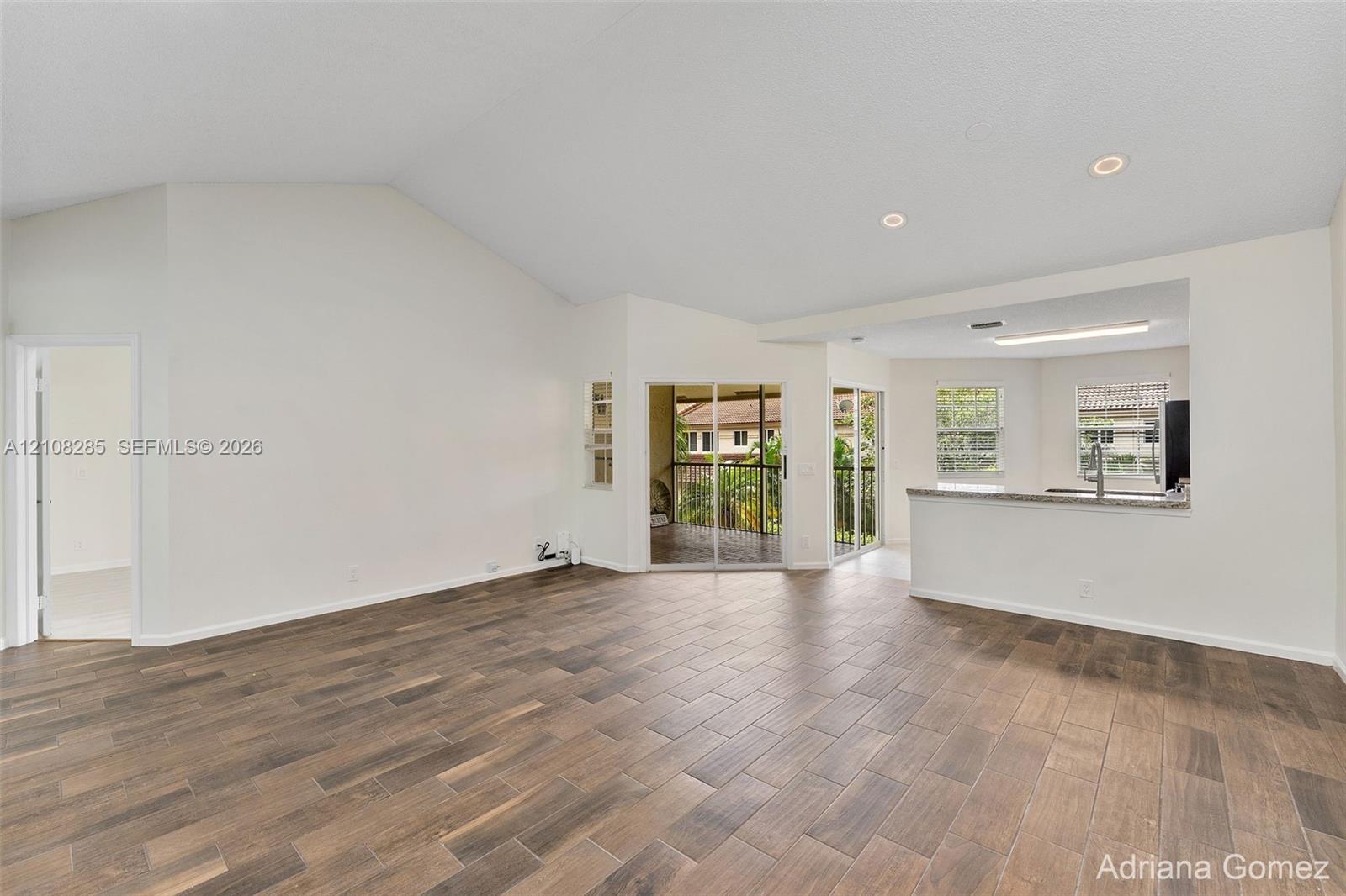 Vaulted ceilings, wood-finish tile flooring, recessed lighting, and sliding glass doors leading to a balcony, complemented by a kitchen pass-through with a stone-top breakfast bar