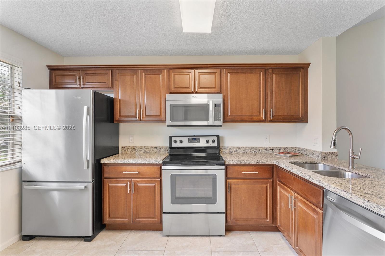Kitchen featuring wooden cabinetry, stone-surface countertops, stainless steel appliances, a double-basin sink with a pull-down faucet, and tiled flooring