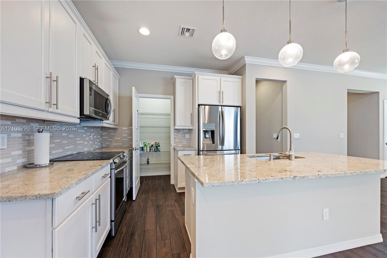 Kitchen featuring white shaker-style cabinetry, stone countertops, stainless steel appliances, wood-finish flooring, and three glass pendant lights over the central island Kitchen featuring white shaker-style cabinetry, stone countertops, stainless steel appliances, wood-finish flooring, and three glass pendant lights over the central island