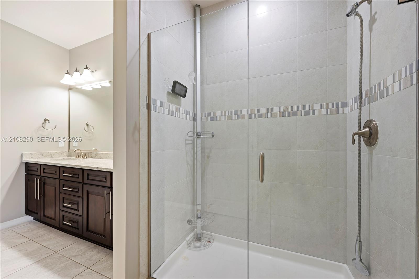 Walk-in shower with a glass enclosure, tile surround, and metallic fixtures, featuring a vanity with stone-finish countertop, dark wood cabinetry, and a full-width wall mirror