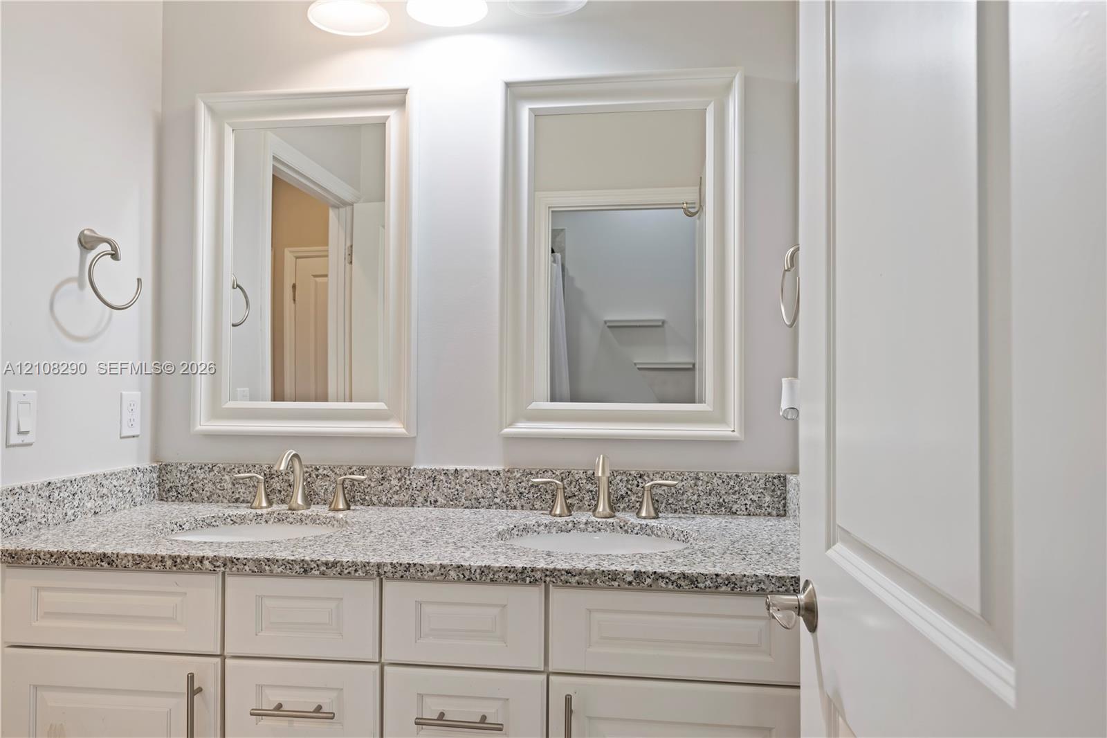 Dual-vanity cabinet featuring white shaker-style cabinetry, speckled stone countertop, integrated undermount sinks, and two framed wall mirrors with three-light fixture overhead