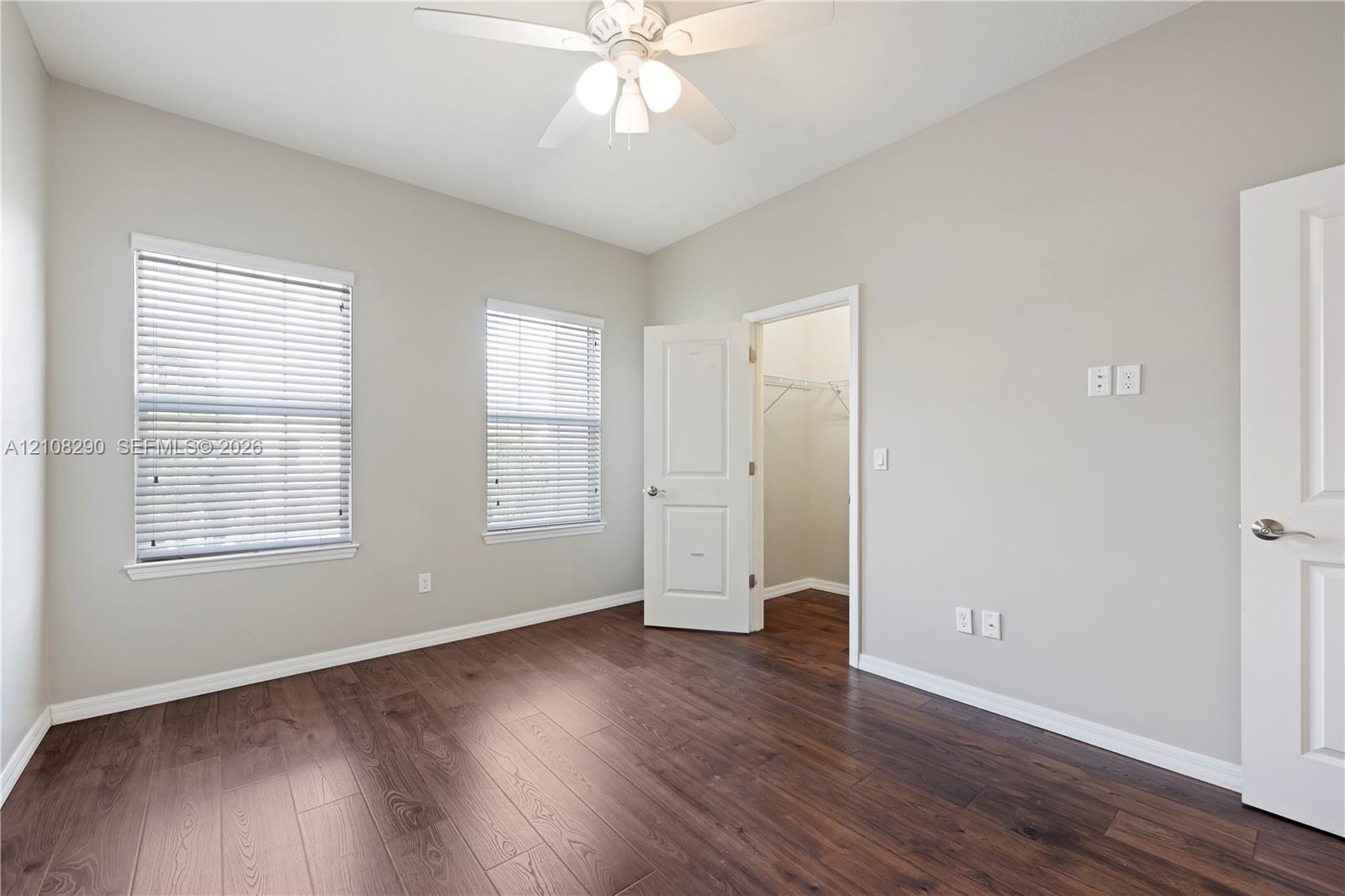 Interior space featuring wood-finish flooring, two windows with horizontal blinds, a ceiling fan with light fixture, and an open doorway to a walk-in closet
