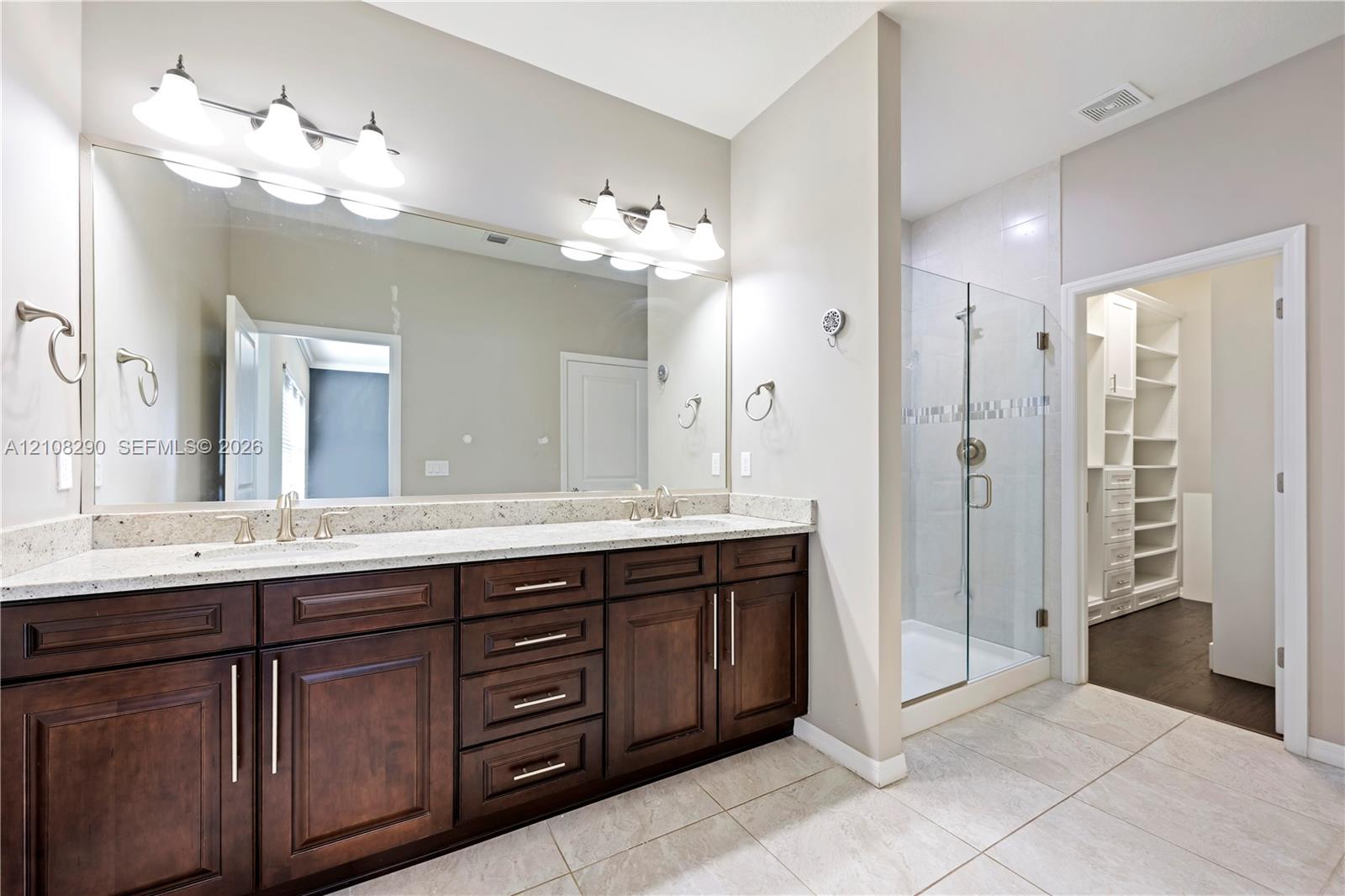 Double vanity featuring dark wood-finish cabinetry, stone-surface countertop, dual undermount sinks, and a large-format vanity mirror with dual light fixtures