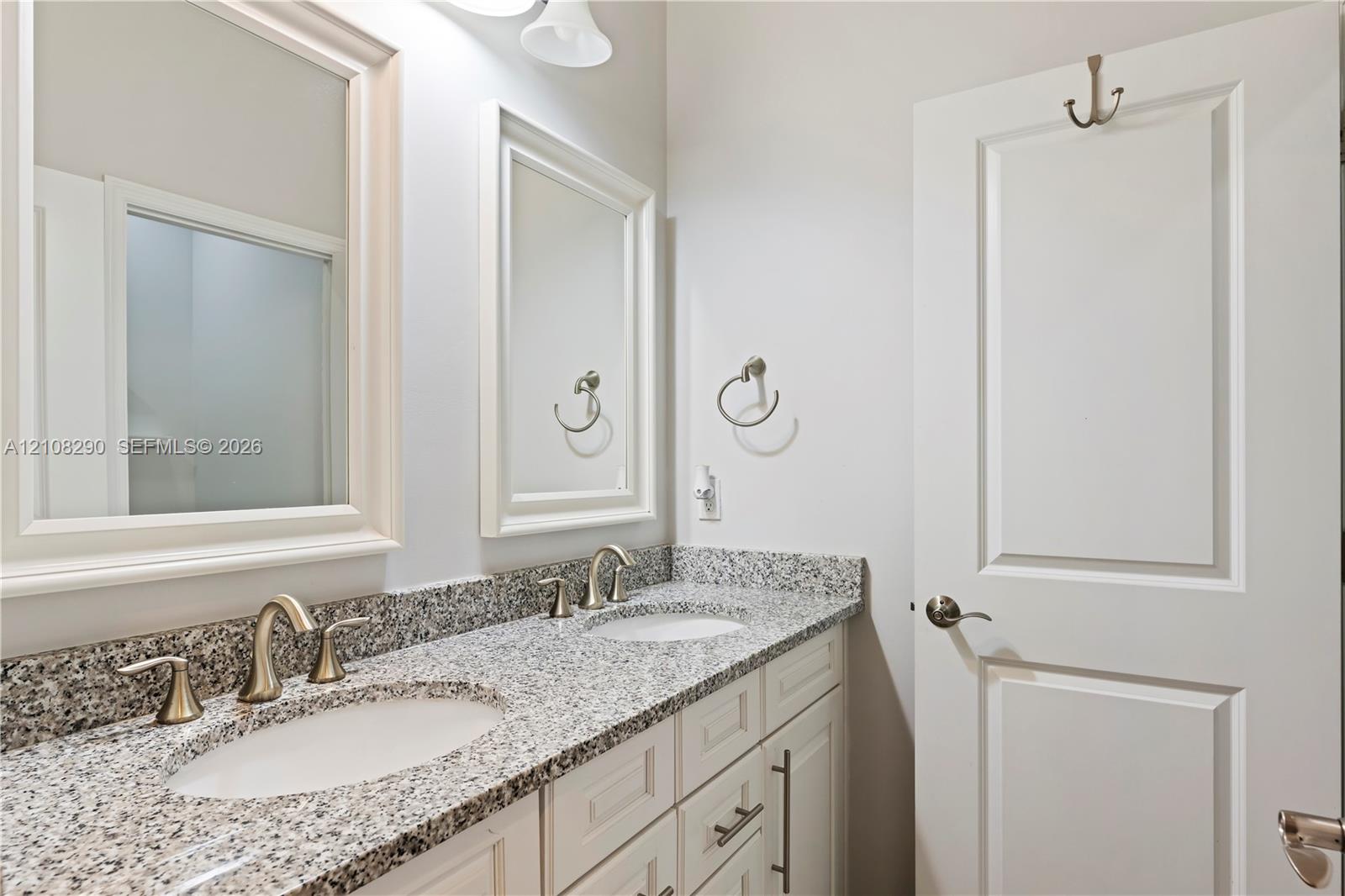 Dual-sink vanity with stone-finish countertop, white shaker-style cabinetry, metallic fixtures, and dual framed wall mirrors