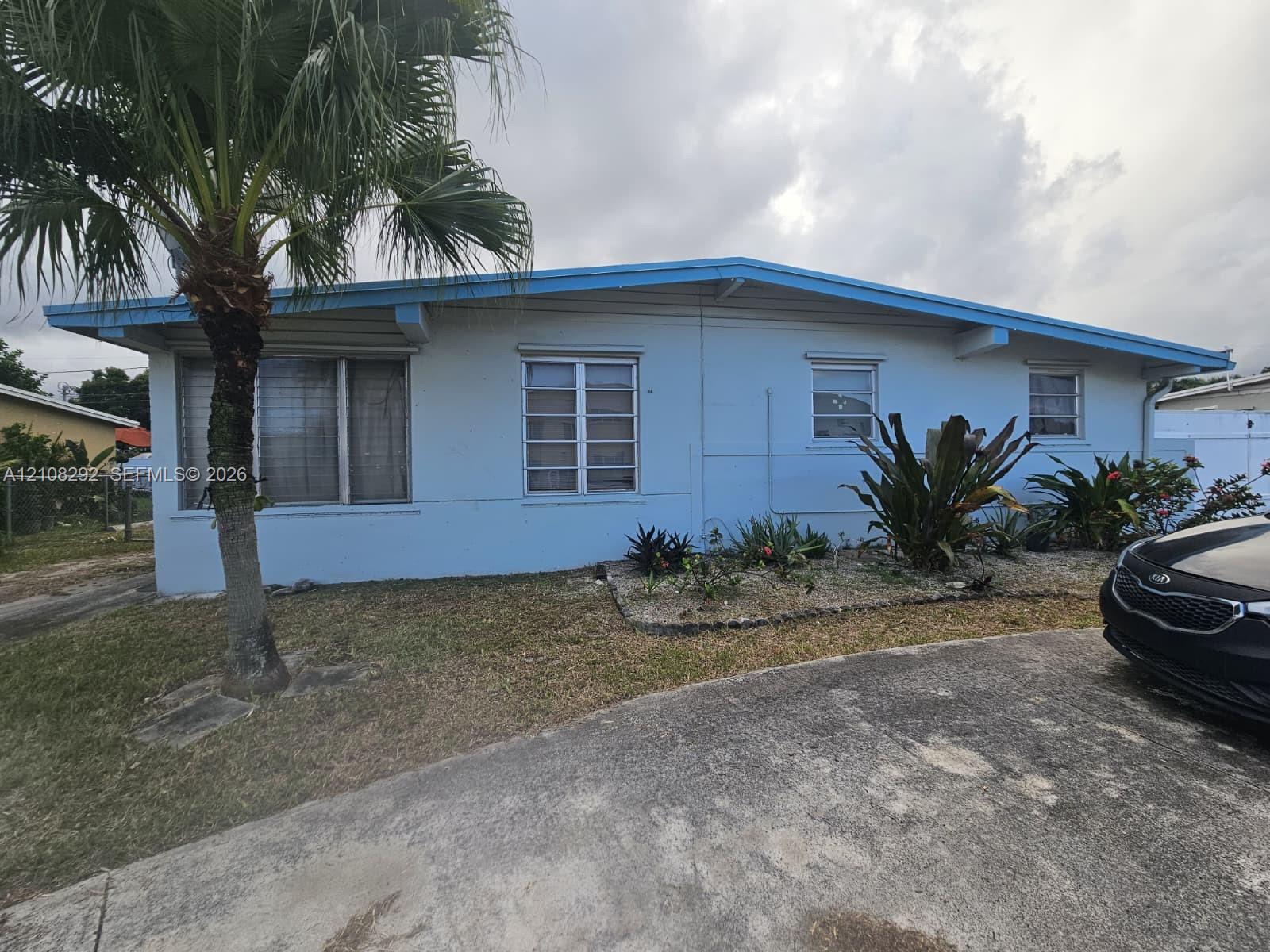 Single-story exterior featuring light blue siding, a low-pitch roofline, exposed rafter tails, metal-frame windows, and a concrete driveway