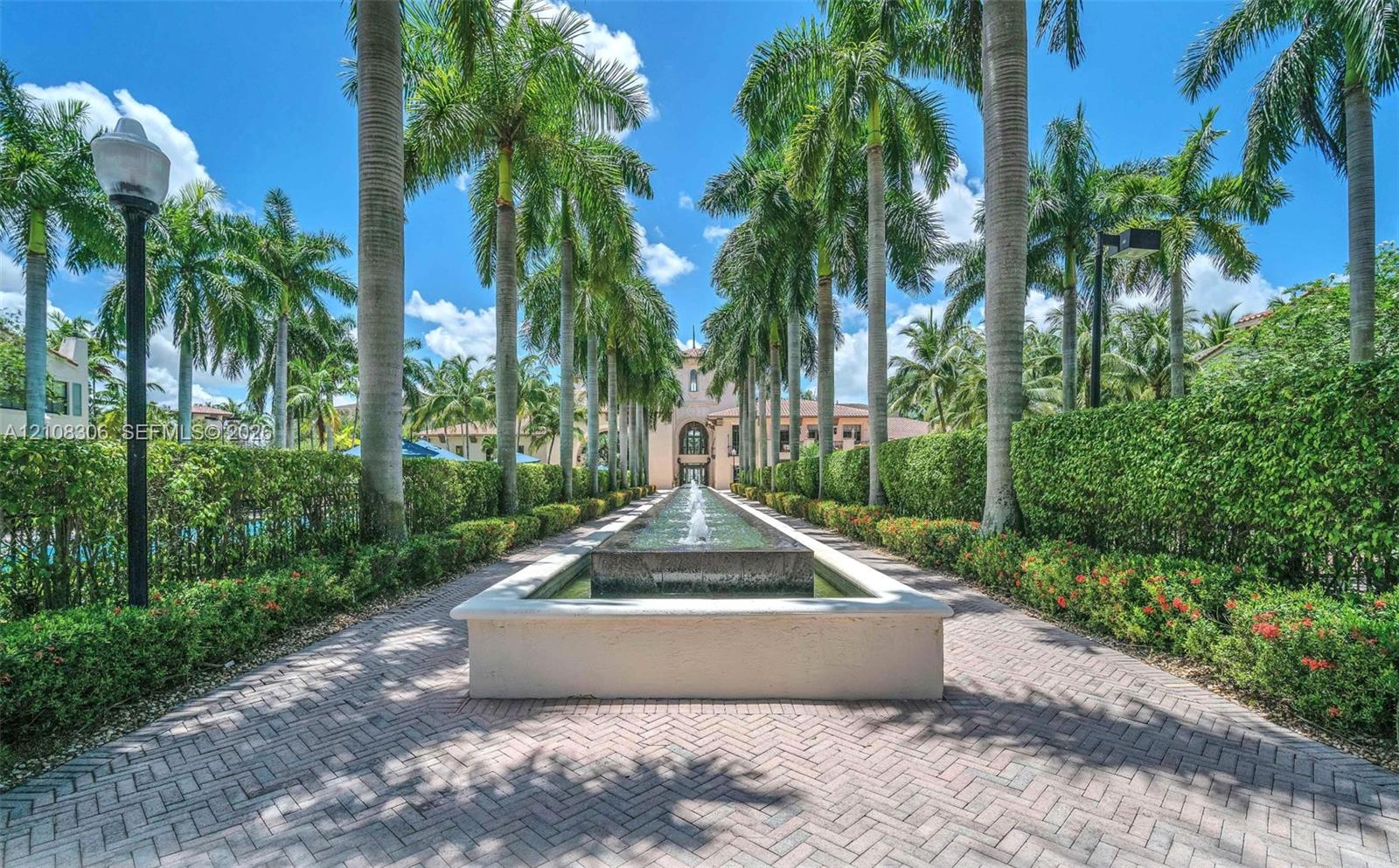 Formal walkway featuring brick-paver flooring, a centered stone-finish fountain, and mature palm trees lined by manicured hedges