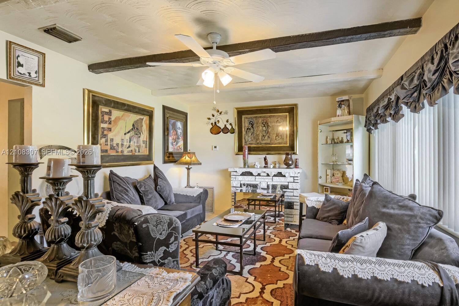 Living area featuring exposed ceiling beams, a white ceiling fan with integrated lighting, and a brick-faced fireplace Living area featuring exposed ceiling beams, a white ceiling fan with integrated lighting, and a brick-faced fireplace