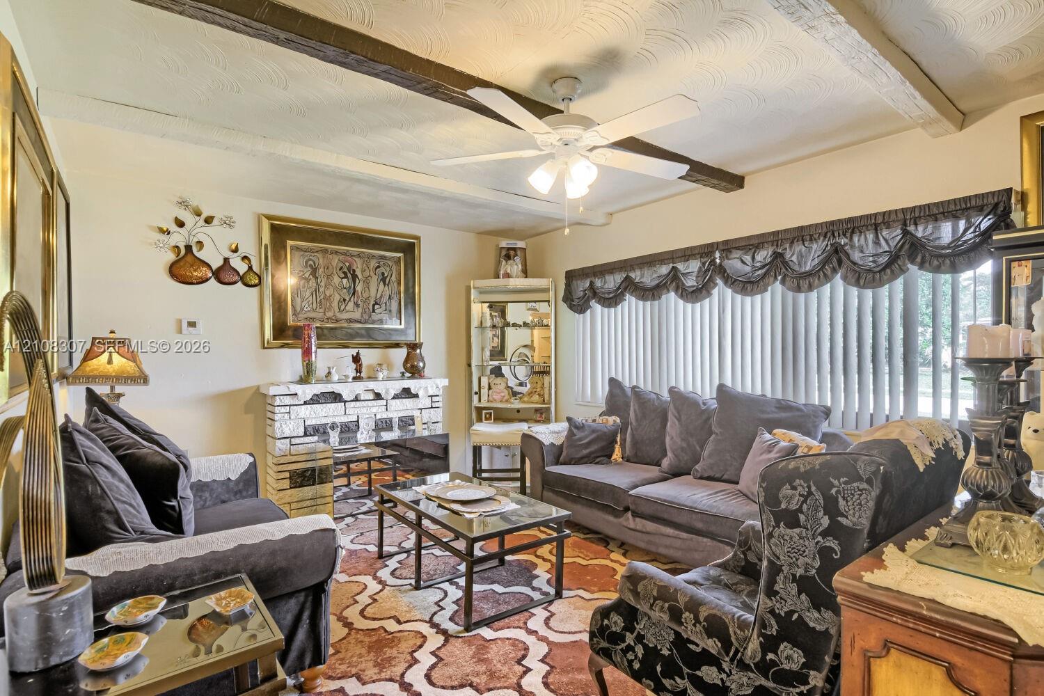 Living area featuring exposed wooden ceiling beams, patterned ceiling detail, stone-faced fireplace, and a large window with horizontal blinds Living area featuring exposed wooden ceiling beams, patterned ceiling detail, stone-faced fireplace, and a large window with horizontal blinds