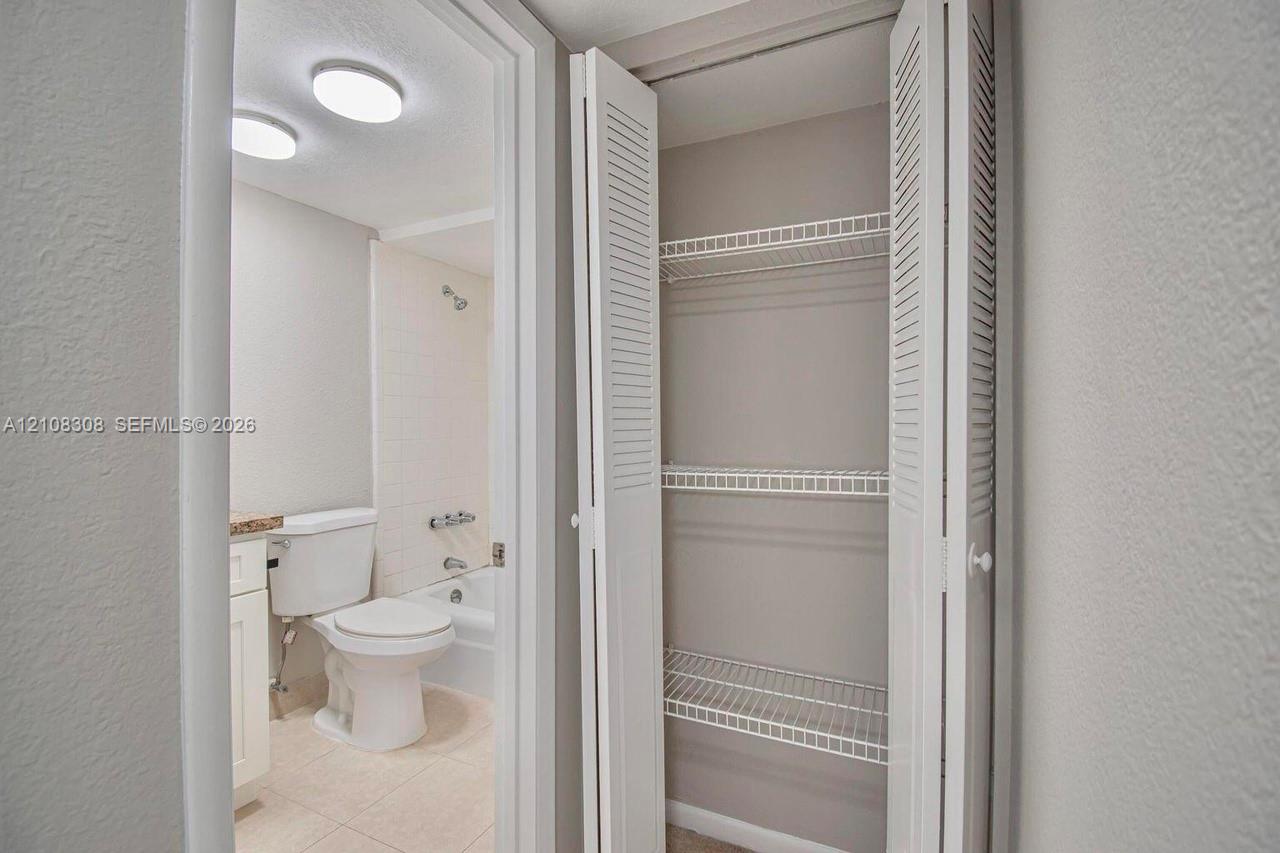 Built-in storage closet with louvered bi-fold doors and wire shelving, featuring a tiled bathtub enclosure, porcelain toilet, and recessed ceiling lighting