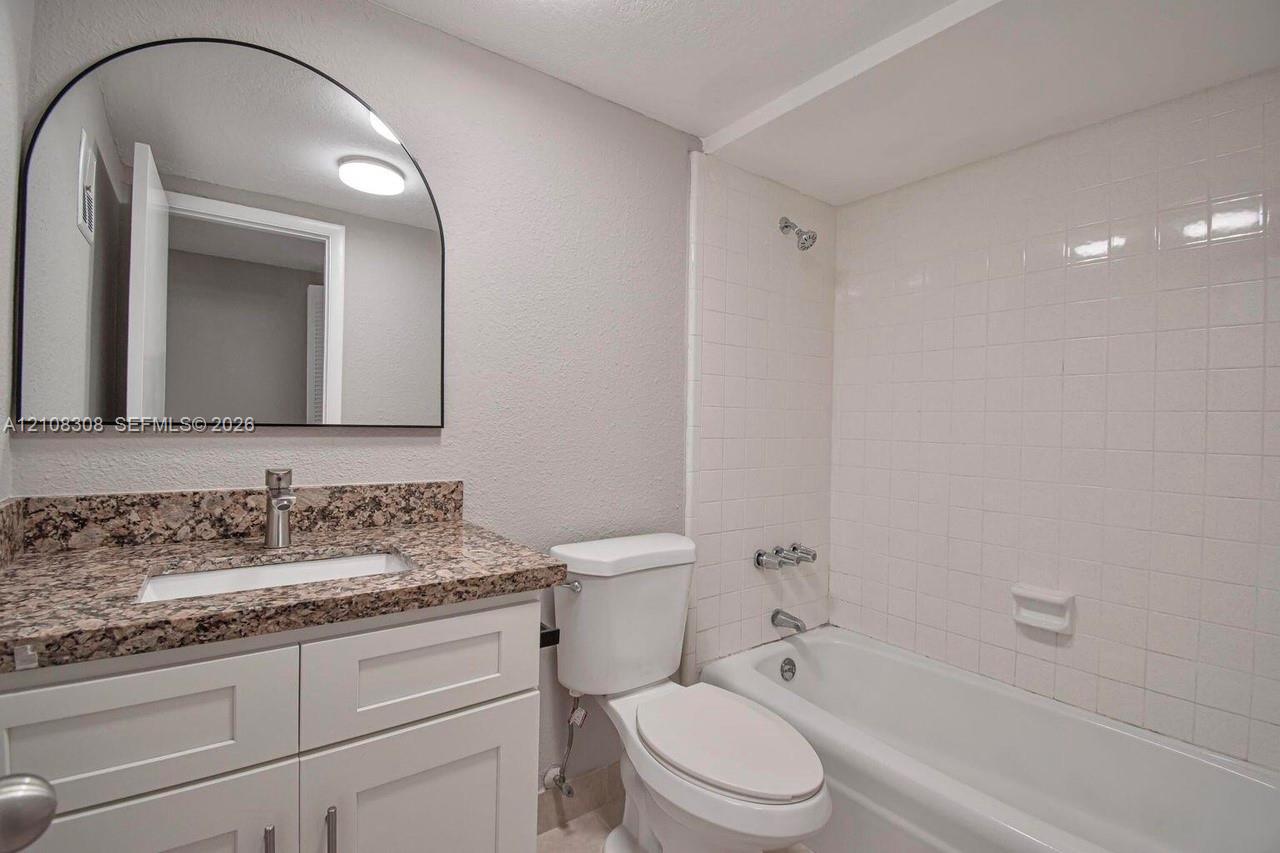 White cabinetry with stone-finish countertop, arched wall mirror, and tiled shower-tub combination