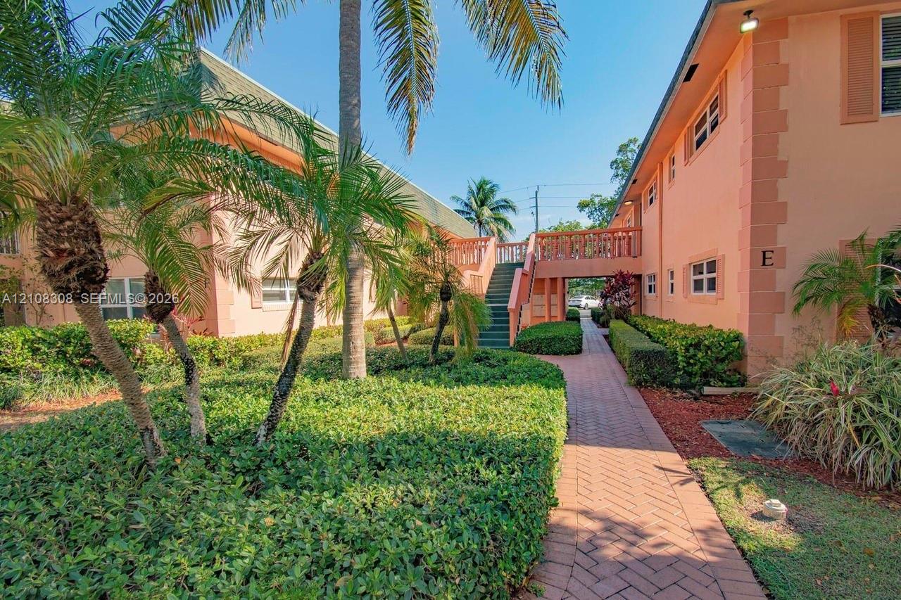 Exterior facade featuring peach-toned stucco, brick-style paver walkways, mature palm trees, sculpted hedges, and a raised exterior staircase with balustrades