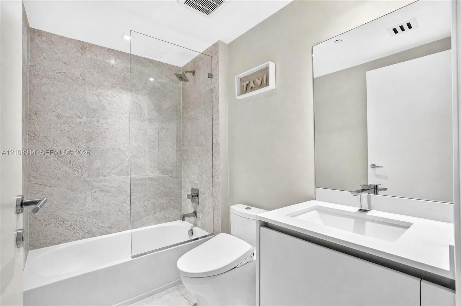 Bathroom featuring a vanity with a white countertop and integrated sink, frameless glass shower enclosure, stone-finish tile surround, and large wall-mounted mirror