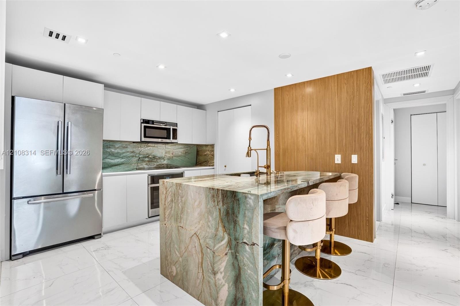 Kitchen featuring flat-panel white cabinetry, stone-clad waterfall island, matching stone backsplash, wood-finish accent wall, and stainless steel appliances Kitchen featuring flat-panel white cabinetry, stone-clad waterfall island, matching stone backsplash, wood-finish accent wall, and stainless steel appliances