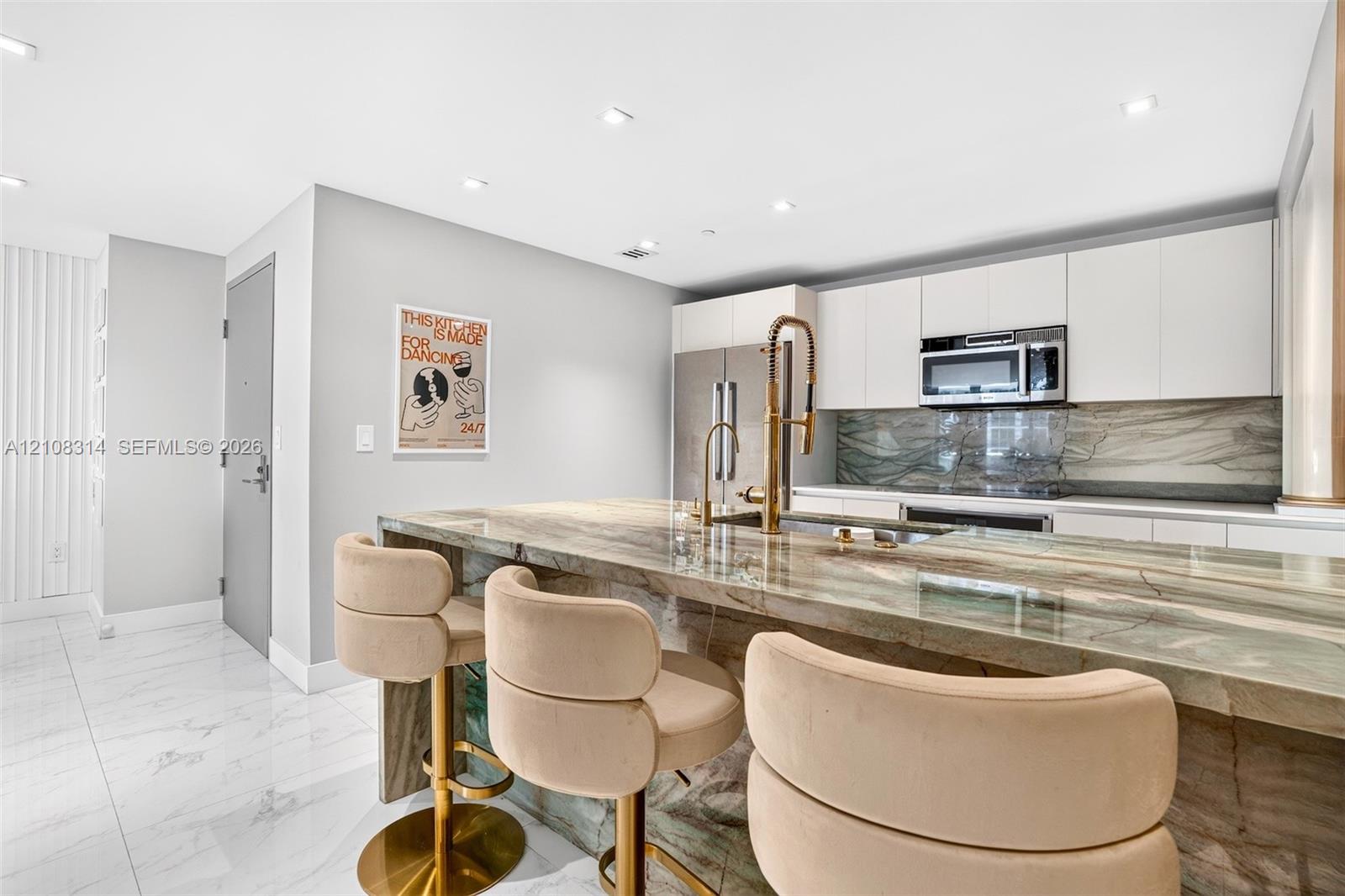 Kitchen featuring a stone waterfall island with a metal gooseneck faucet, white flat-panel cabinetry, a matching stone backsplash, stainless steel appliances, and large-format tile flooring Kitchen featuring a stone waterfall island with a metal gooseneck faucet, white flat-panel cabinetry, a matching stone backsplash, stainless steel appliances, and large-format tile flooring