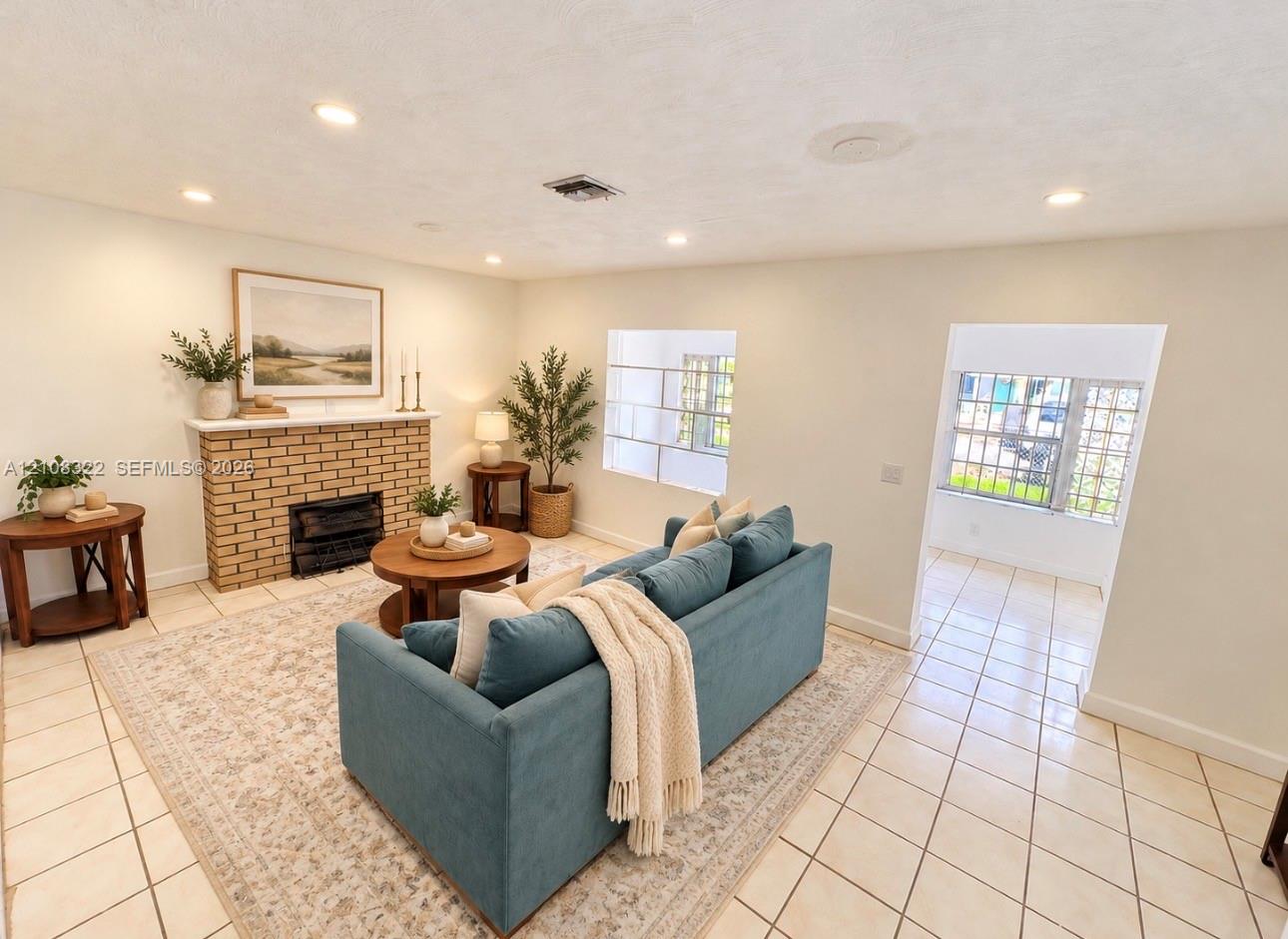 Living area featuring light-toned tile flooring, a brick masonry fireplace with a mantel, recessed ceiling lighting, and windows with decorative geometric grilles