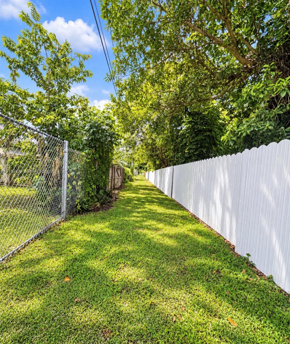 Grassy exterior area featuring a white scalloped-top vinyl fence, chain-link perimeter fencing, and mature trees