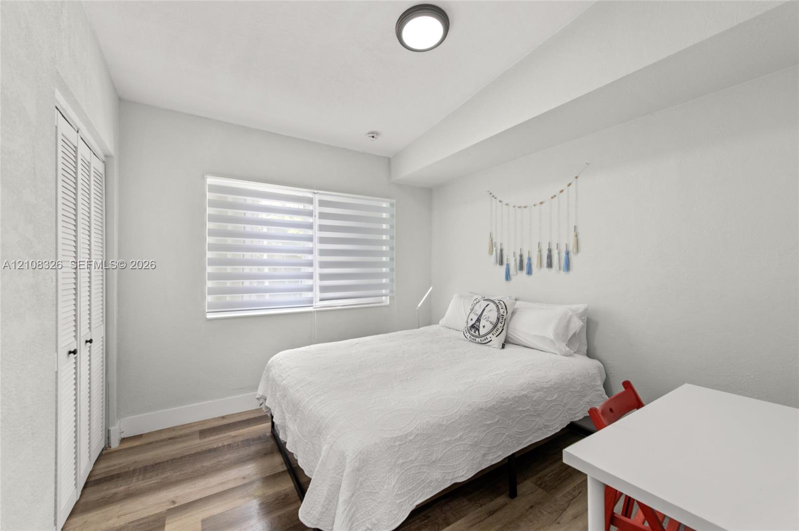 Neutral-toned room featuring wood-finish flooring, dual-pane windows with adjustable roller shades, and floor-to-ceiling closet with louvered bi-fold doors Neutral-toned room featuring wood-finish flooring, dual-pane windows with adjustable roller shades, and floor-to-ceiling closet with louvered bi-fold doors