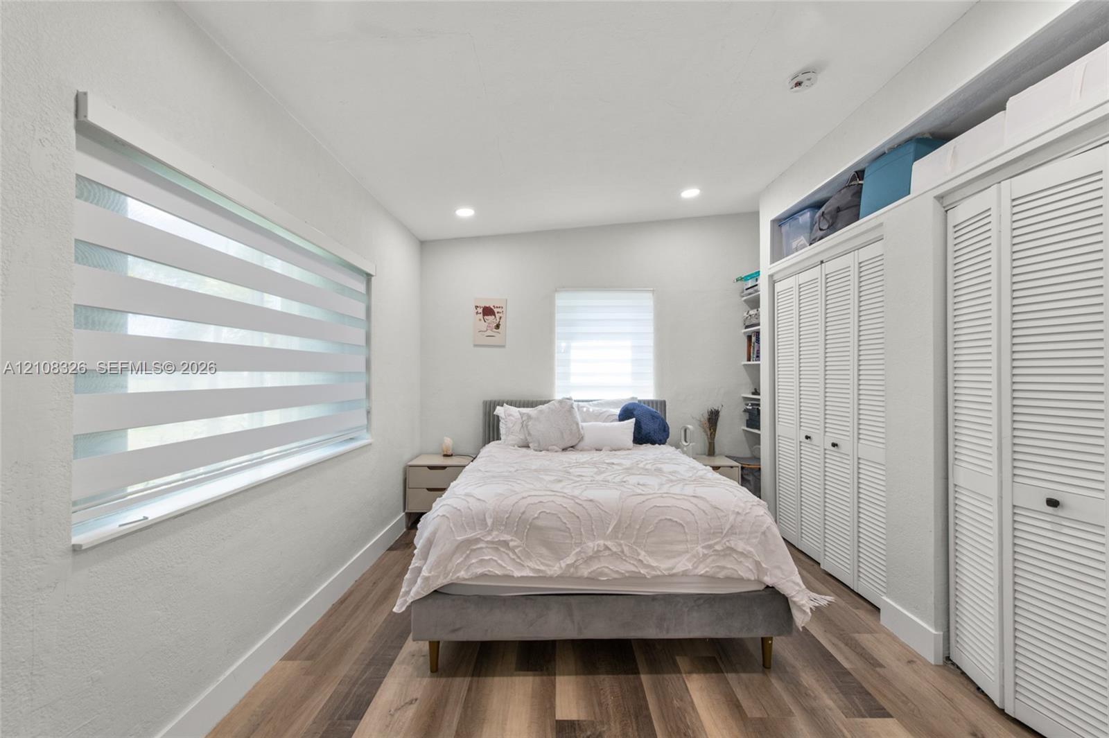 Wood-finish flooring, recessed lighting, dual-window configuration with zebra shades, and floor-to-ceiling louvered closet doors with overhead storage Wood-finish flooring, recessed lighting, dual-window configuration with zebra shades, and floor-to-ceiling louvered closet doors with overhead storage