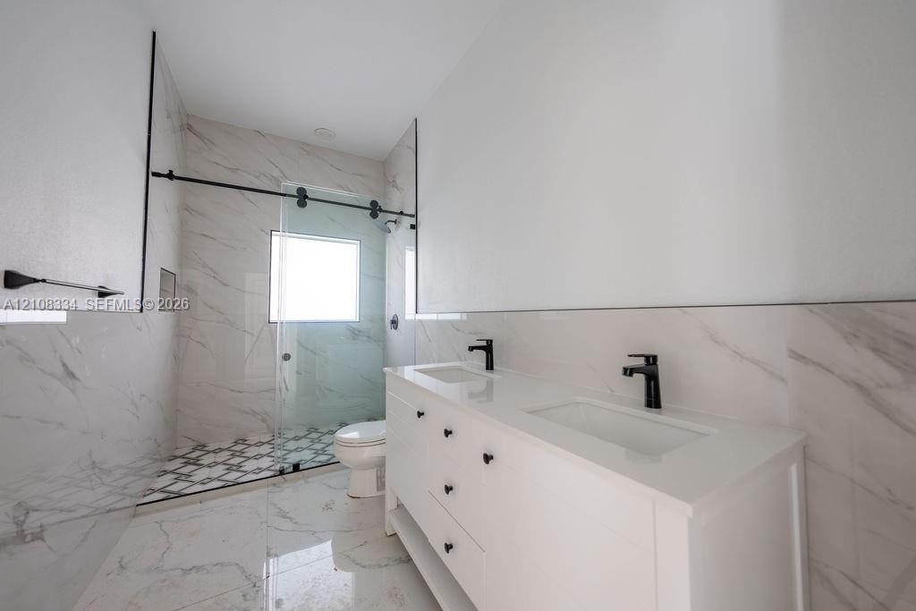 Dual-sink vanity with a white countertop and matte black fixtures, walk-in shower with a glass sliding door, large-format stone-patterned tile flooring, and floor-to-ceiling tiled shower surround