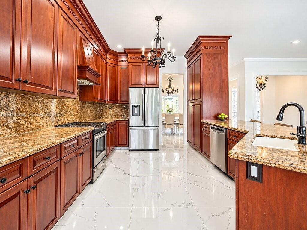 Kitchen featuring custom cherry wood cabinetry with crown molding, stone countertops, stainless steel appliances, polished tile flooring with veining, and a central decorative chandelier