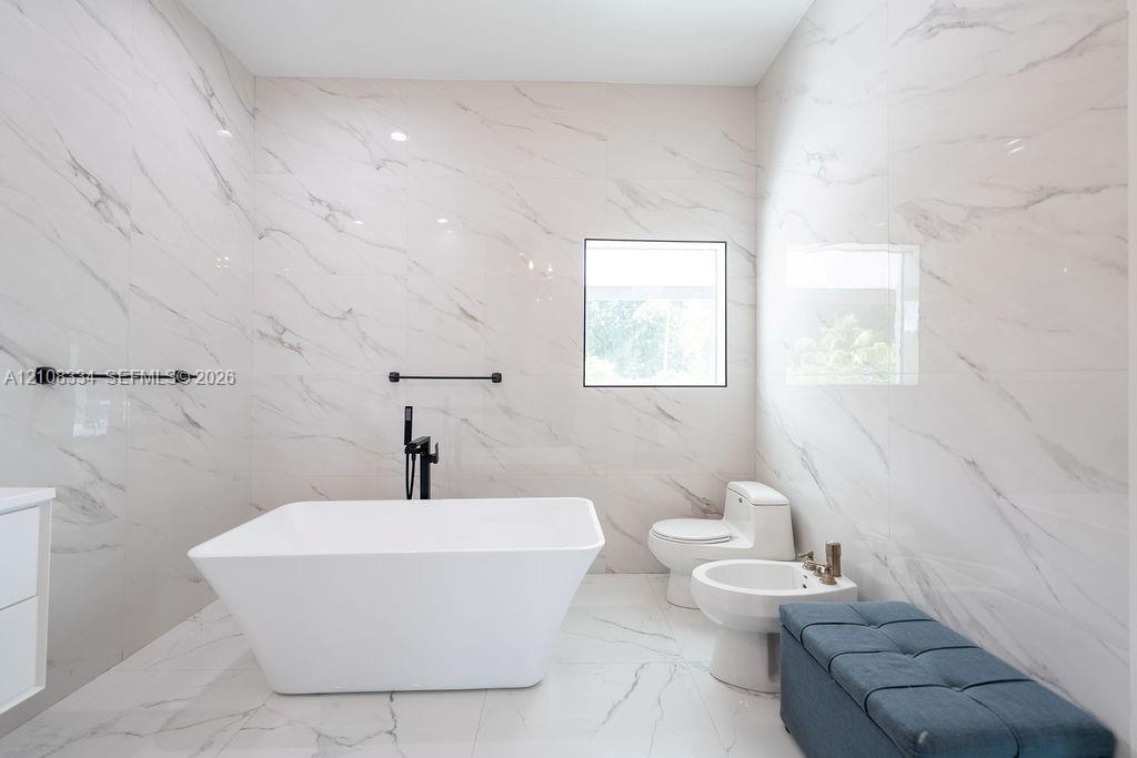 Floor-to-ceiling porcelain tile with veining, freestanding rectangular soaking tub, matte black wall-mounted hardware, and square window