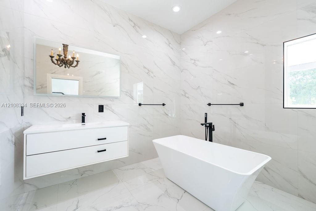 Floor-to-ceiling stone-style wall and floor tile, white floating dual-drawer vanity, freestanding soaking tub, matte black wall-mounted fixtures, and a rectangular mirror