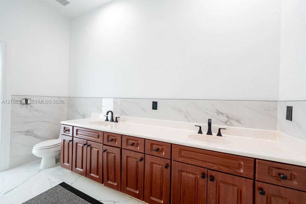 Dual-vanity cabinetry with wood finish, integrated stone-surface countertop, dual undermount sinks, dark metal hardware, and large-format tile wall backsplash