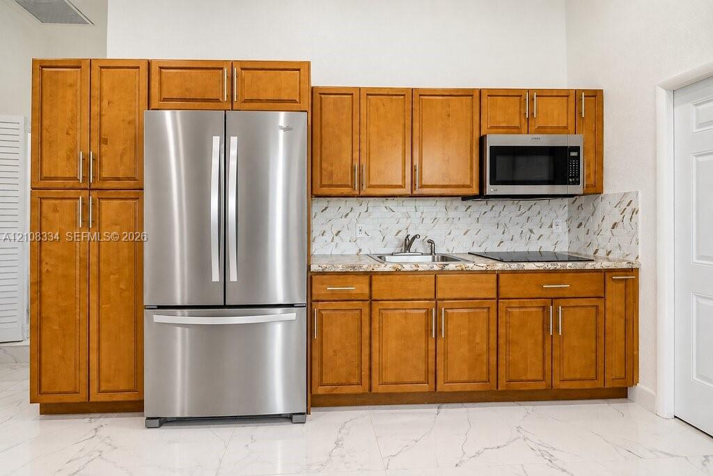 Kitchenette featuring shaker-style wood cabinetry, stainless steel refrigerator and built-in microwave, patterned backsplash, and stone-finish countertop