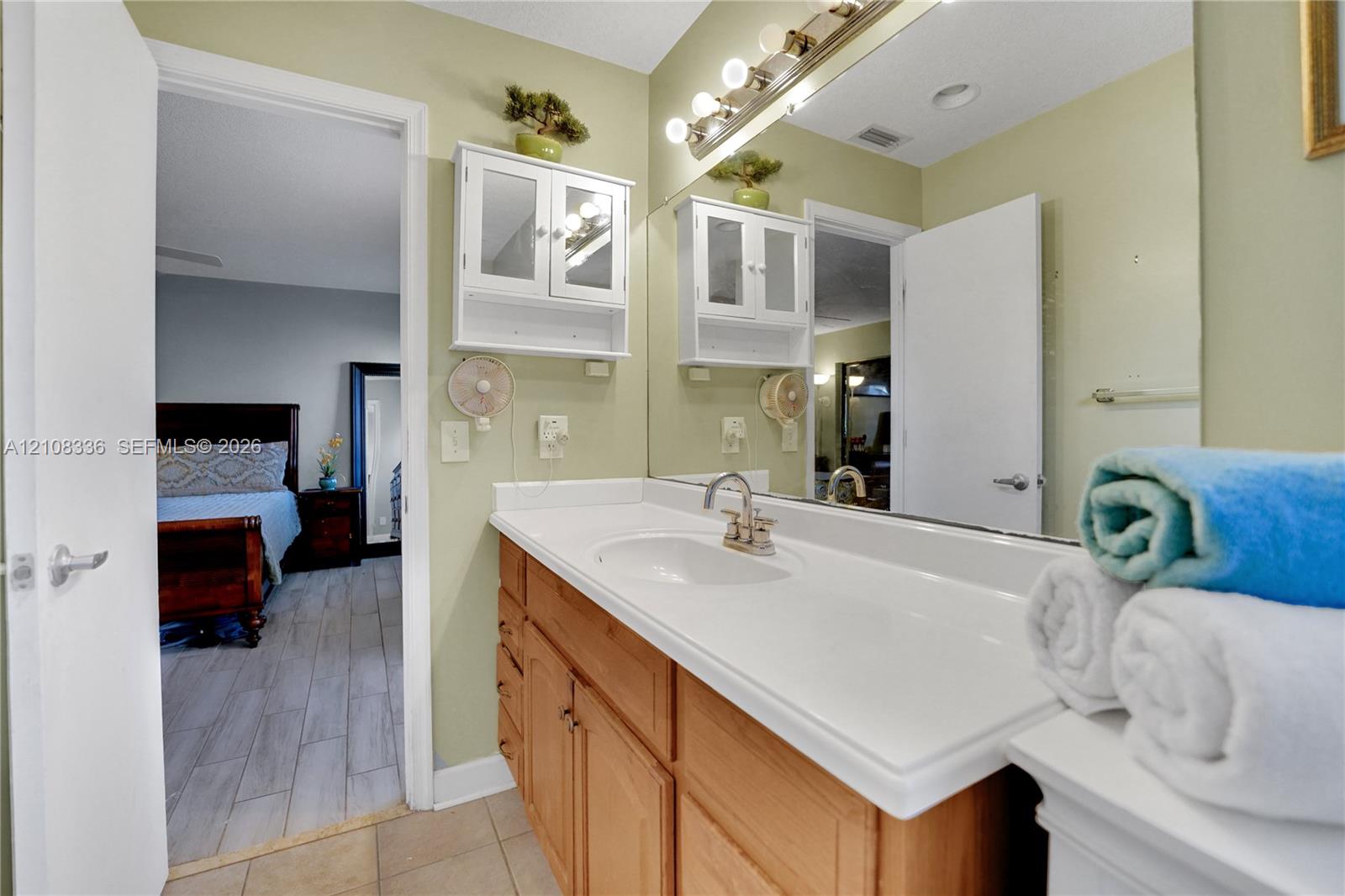 Single-sink vanity with a solid-surface countertop, natural wood-finish cabinetry, wall-mounted white storage hutch, and large-scale vanity mirror with integrated multi-bulb lighting