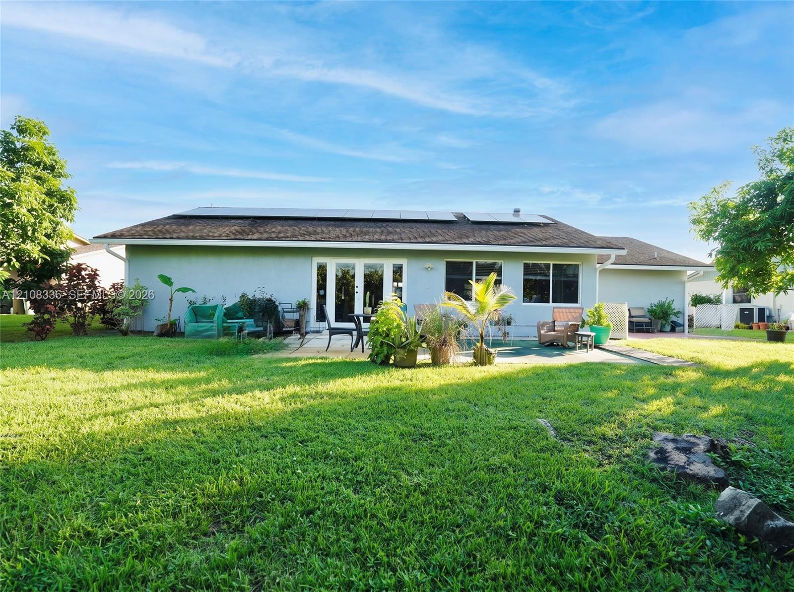 Exterior rear elevation featuring a stucco facade, rooftop solar panel array, multi-panel glass door, and a paved patio area set within a spacious lawn