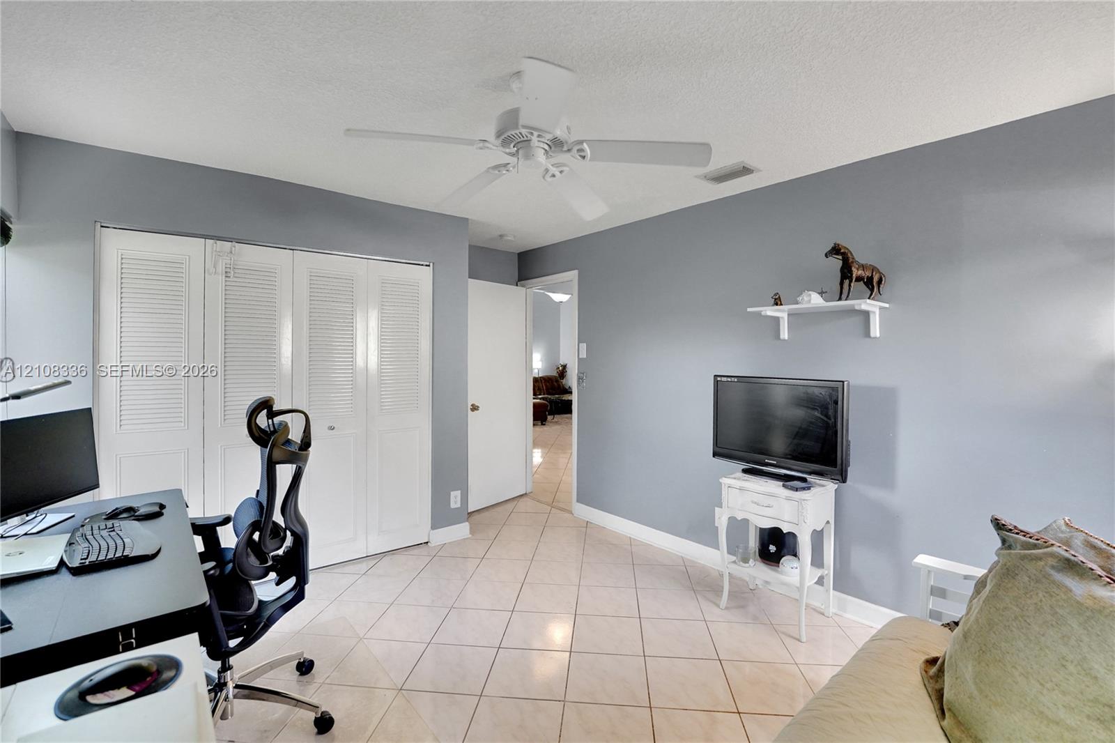 Room featuring neutral-toned walls, tile flooring, louvered bi-fold closet doors, a white ceiling fan, and a wall-mounted display shelf