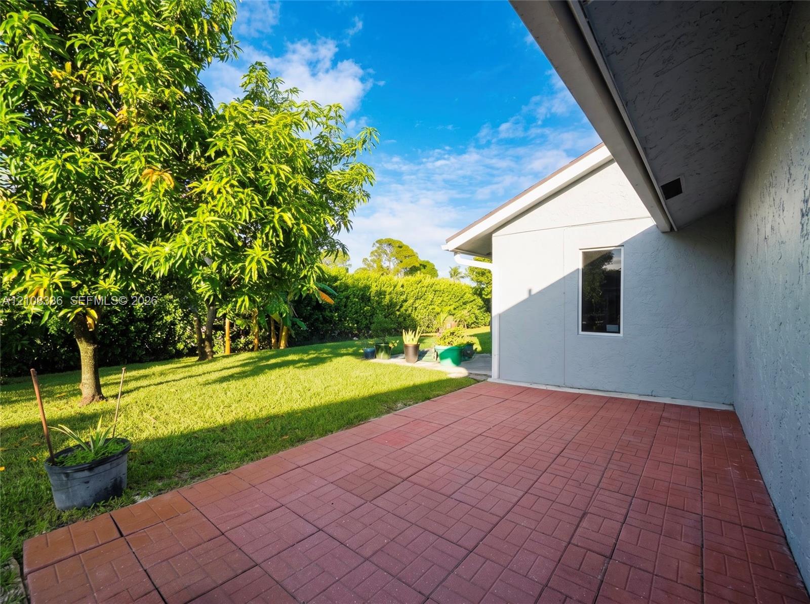 Rear exterior featuring a red brick-patterned paved patio, stucco siding, and a landscaped backyard with mature trees