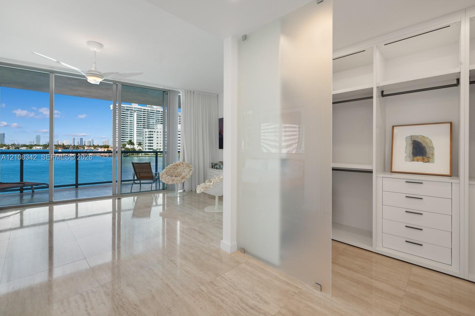 Floor-to-ceiling glass wall with sliding doors, polished stone tile flooring, frosted glass privacy panel, custom built-in wardrobe with soft-close drawers, and integrated ceiling fan with lighting