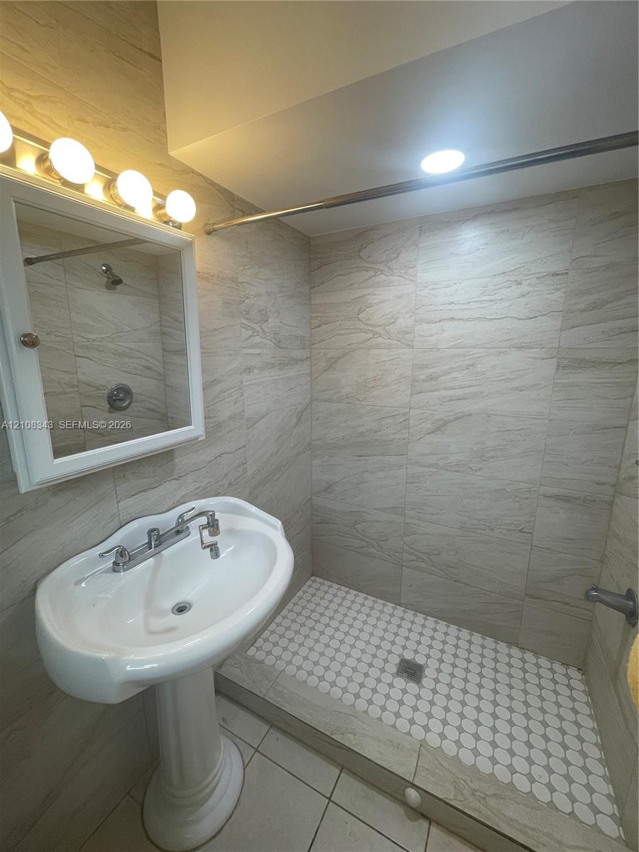 Bathroom featuring floor-to-ceiling neutral stone-finish tile, walk-in shower with penny-round floor tile, pedestal sink with chrome faucet, and mirrored medicine cabinet with integrated four-light fixture
