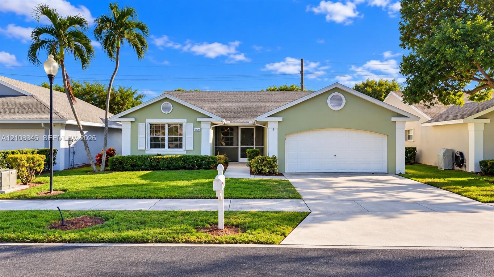 Single-story exterior featuring a gabled roofline, light green siding with white trim, a multi-car garage, and a manicured lawn with mature palm trees