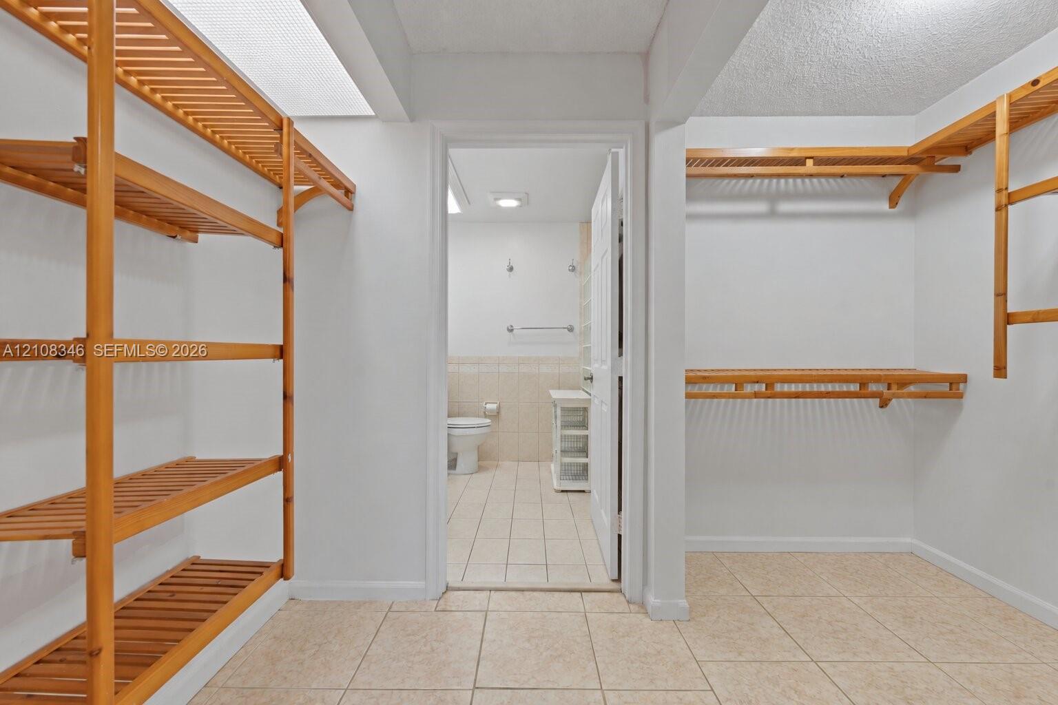Walk-in closet featuring light-toned tile flooring and custom wood slatted shelving, with entry to a bathroom showcasing tile wainscoting and a white vanity cabinet