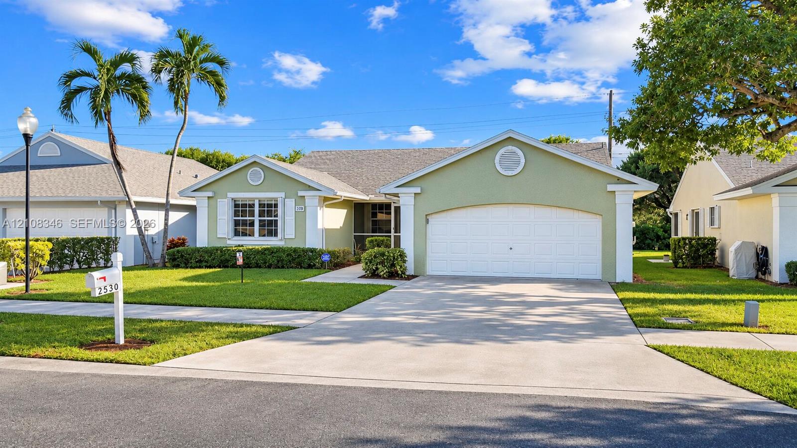 Single-story residence featuring a light green stucco exterior, an arched multi-car garage door, decorative gable vents, and a concrete driveway Single-story residence featuring a light green stucco exterior, an arched multi-car garage door, decorative gable vents, and a concrete driveway