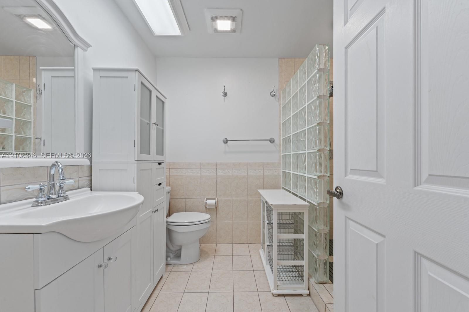 Bathroom featuring a white vanity cabinet with an integrated sink, ceramic tile flooring, neutral wall-to-wall ceramic tiling, and a glass block privacy partition