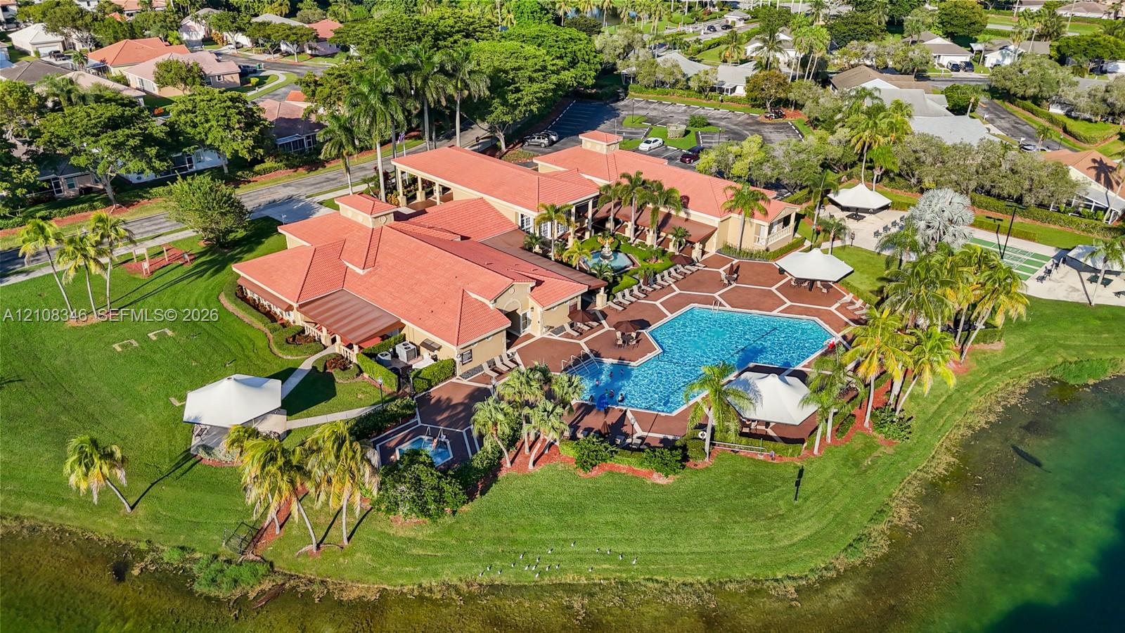 Aerial view of a clubhouse in the community center featuring a tile-roofed structure, a large swimming pool, an attached spa, paved patio decking, and landscaped grounds