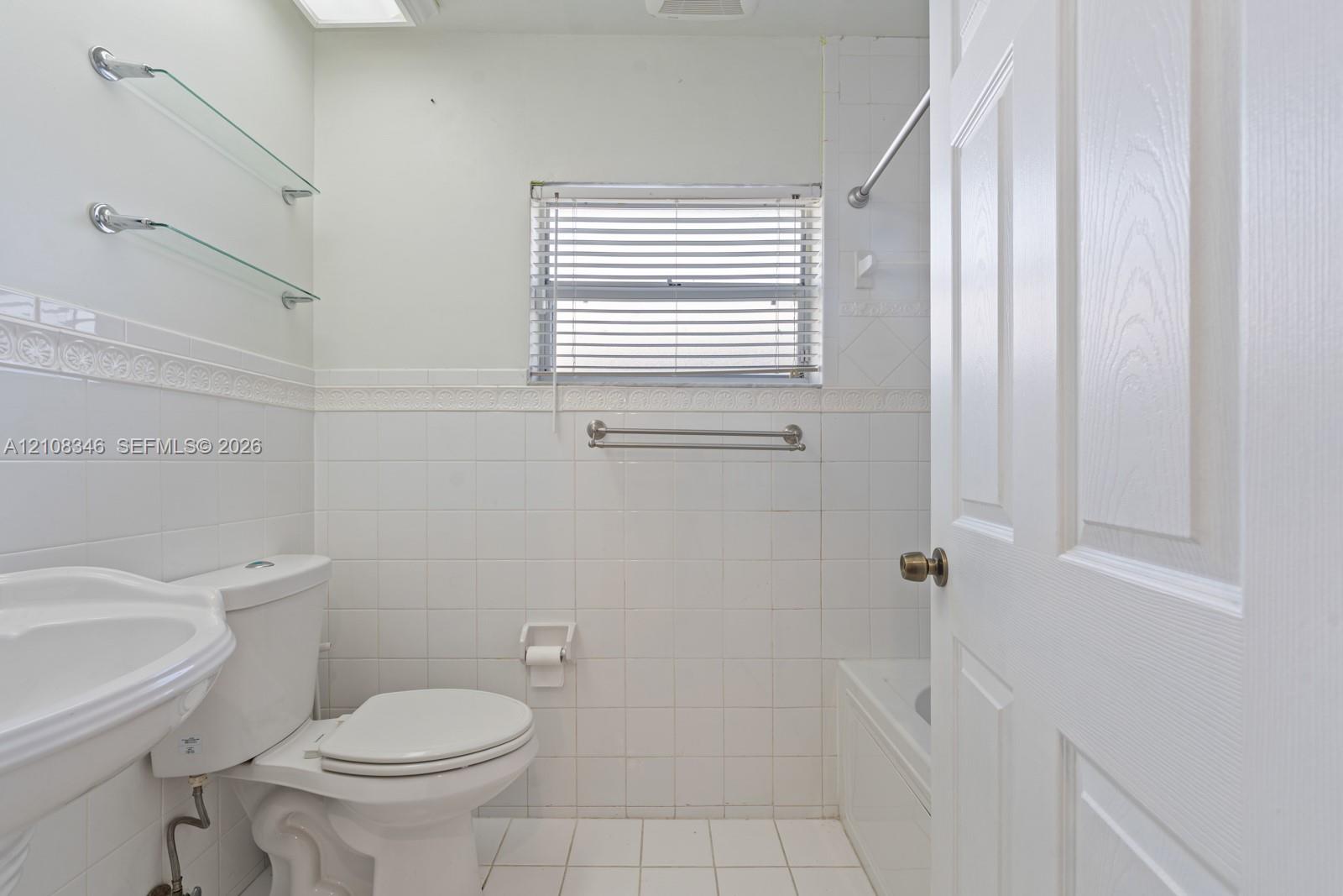Bathroom featuring white ceramic wall and floor tiling, a pedestal sink, a built-in bathtub with a metal shower rod, and two integrated glass shelving units