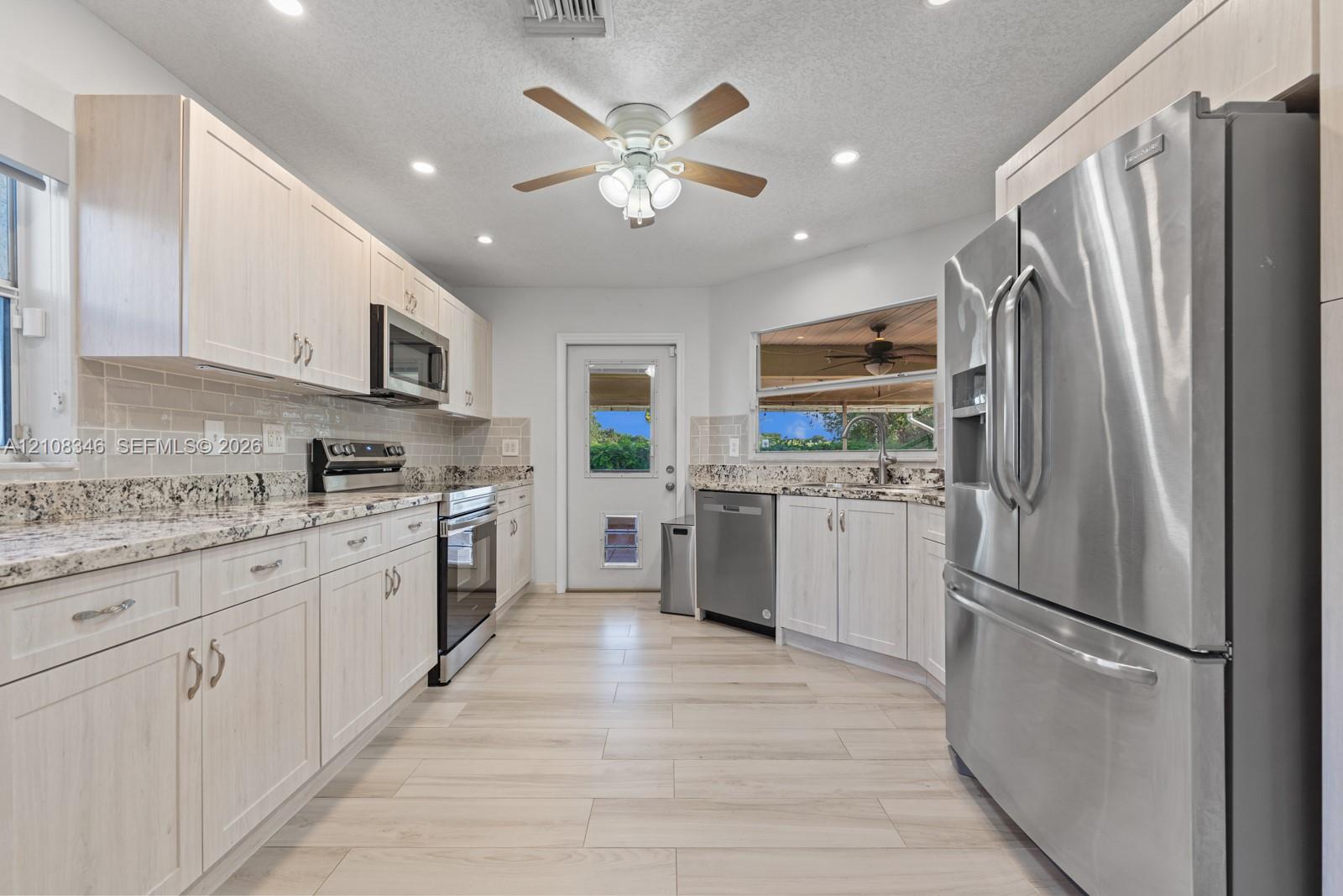 Kitchen featuring light-toned cabinetry, stone-finish countertops, stainless steel appliances, tile backsplash, and wood-tile flooring and exterior door to lani Kitchen featuring light-toned cabinetry, stone-finish countertops, stainless steel appliances, tile backsplash, and wood-tile flooring and exterior door to lani