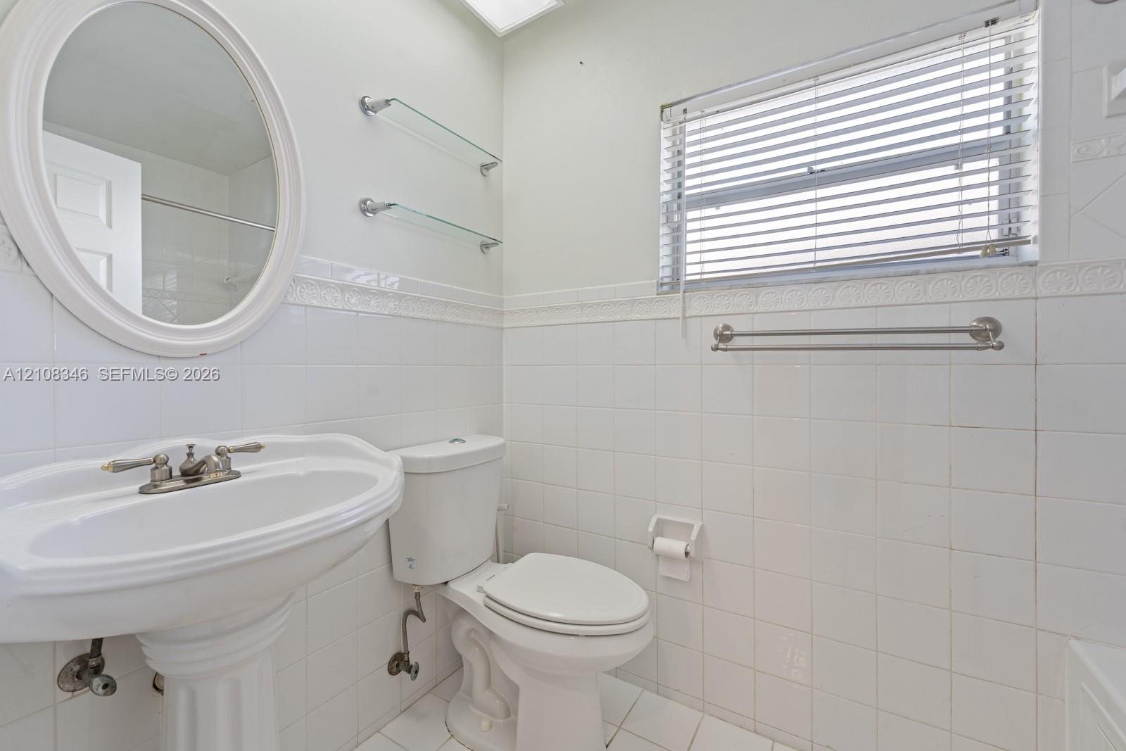 Bathroom featuring white ceramic tile wall cladding, pedestal sink with polished metal faucet, oval wall-mounted mirror, glass shelving, and horizontal window blinds