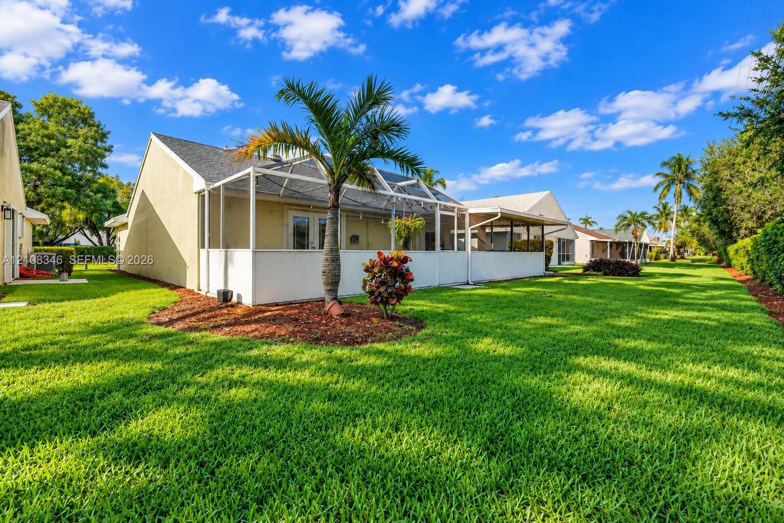 Rear exterior featuring a screened-in lanai, manicured lawn, palm trees, and ground-level plantings