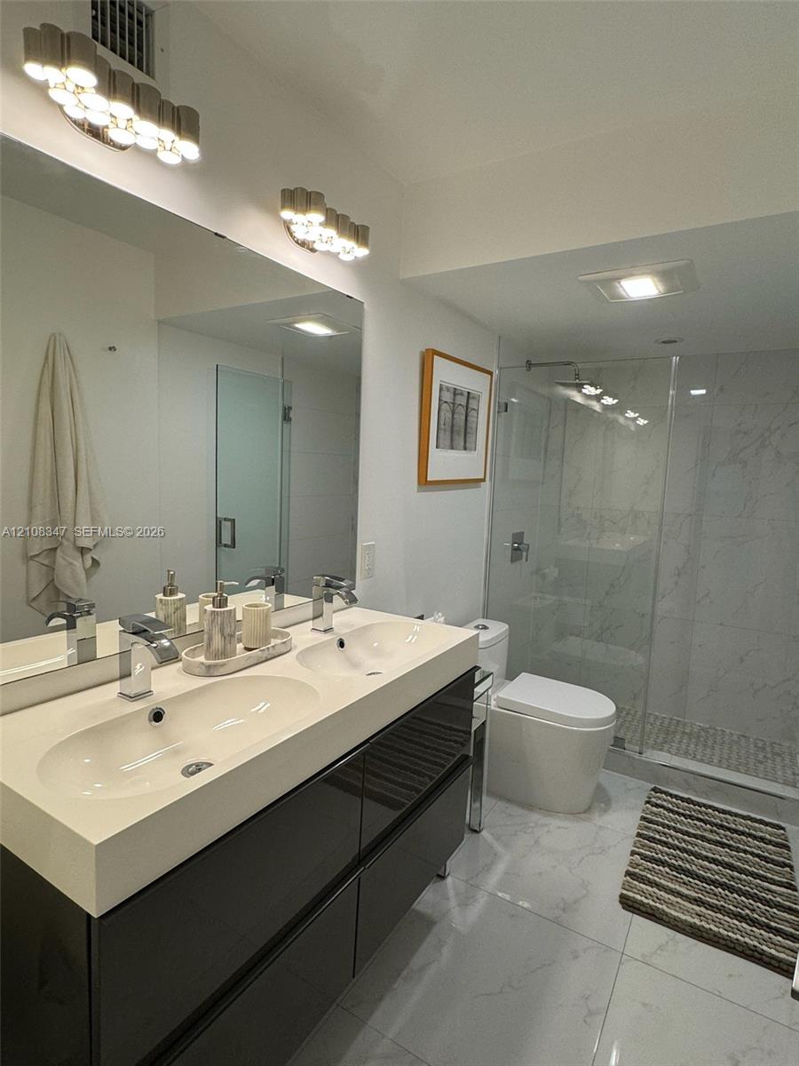 Dual-sink vanity with high-gloss cabinetry and integrated countertop, glass-enclosed walk-in shower with tiled surround, and polished large-format floor tiling
