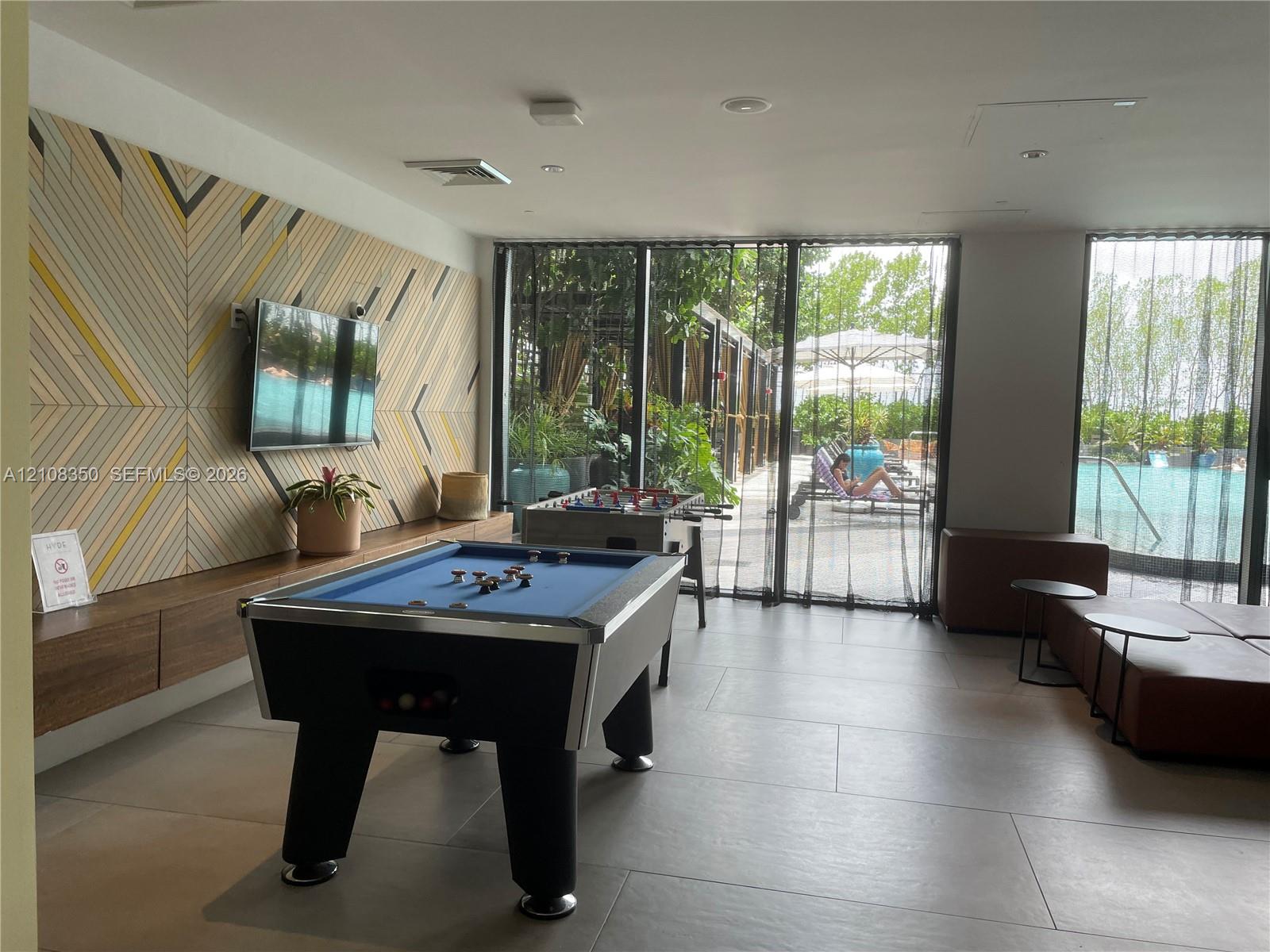Recreation room featuring large-format porcelain tile flooring, geometric accent wall paneling, built-in floating wood cabinetry, and floor-to-ceiling glass panel windows